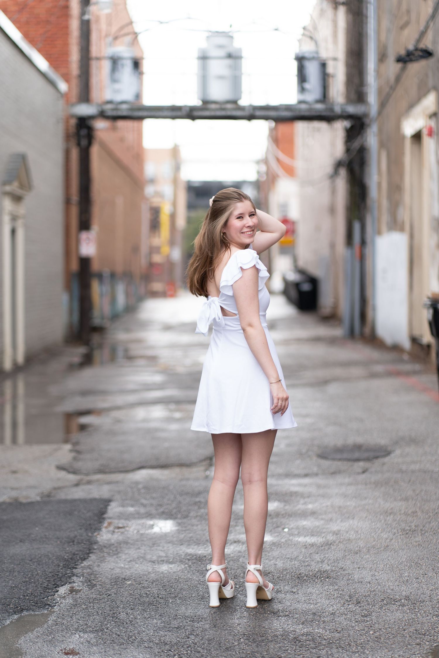 A person in a white summer dress and metallic heels poses in an urban alley with industrial pipes overhead.