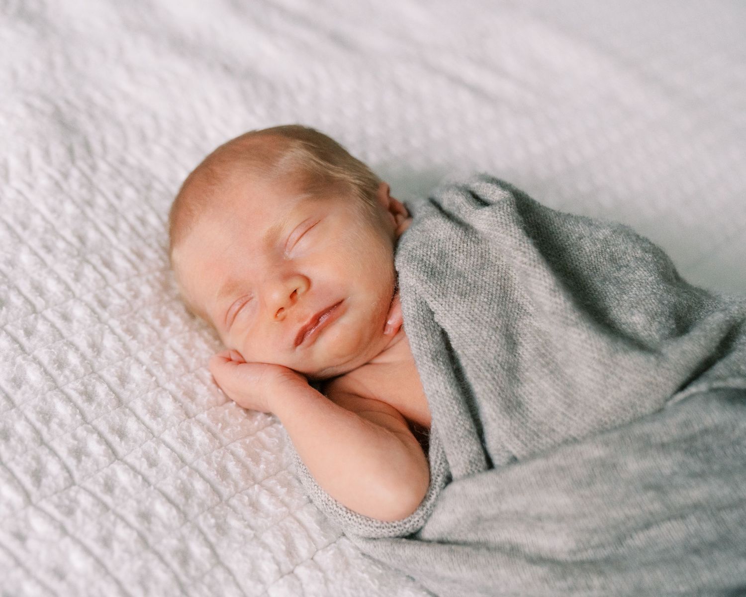Peaceful newborn baby sleeping while wrapped in soft gray blanket on white bedding.