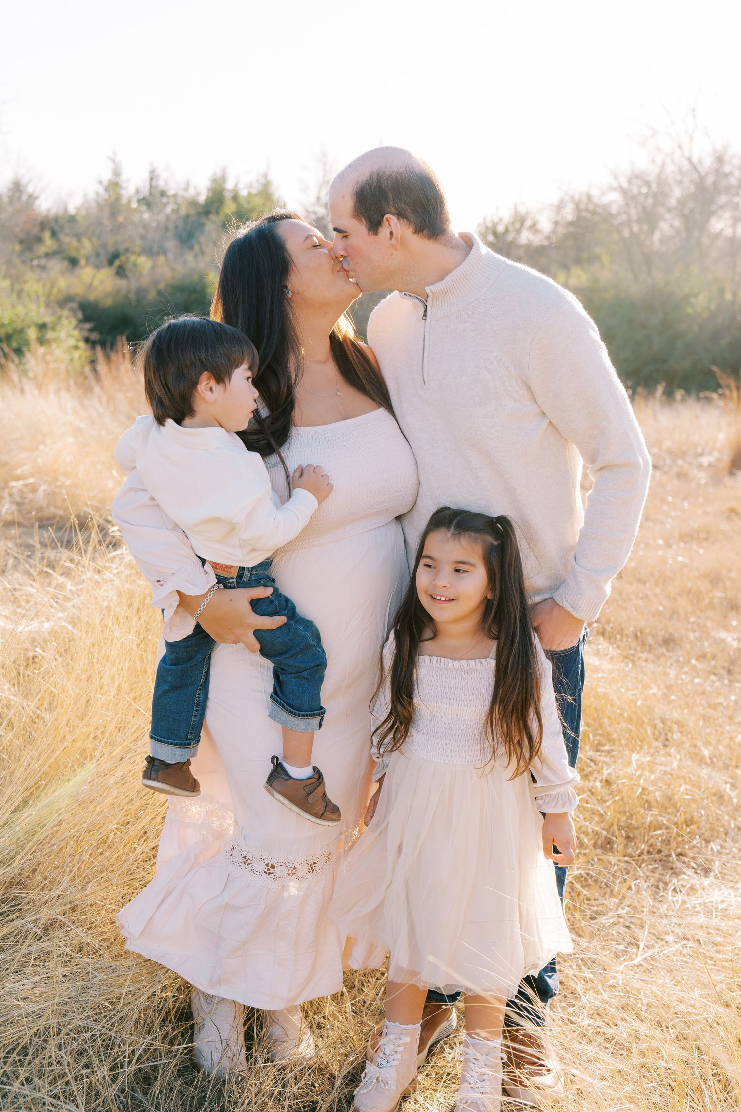 A family wearing white outfits poses together in a sunlit field of tall golden grass during sunset.
