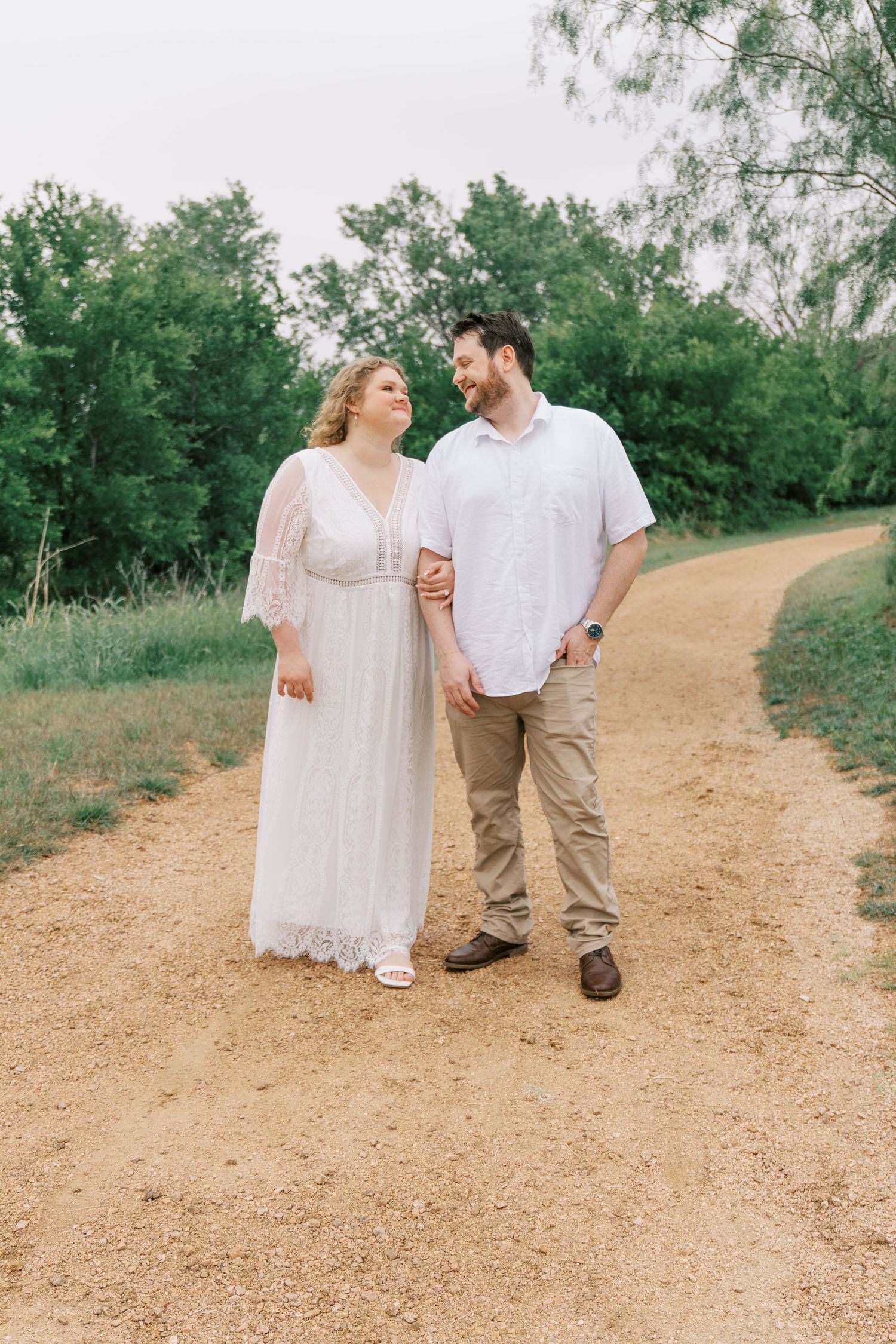 Couple in white outfits holding hands and smiling at each other while walking on a scenic dirt path.