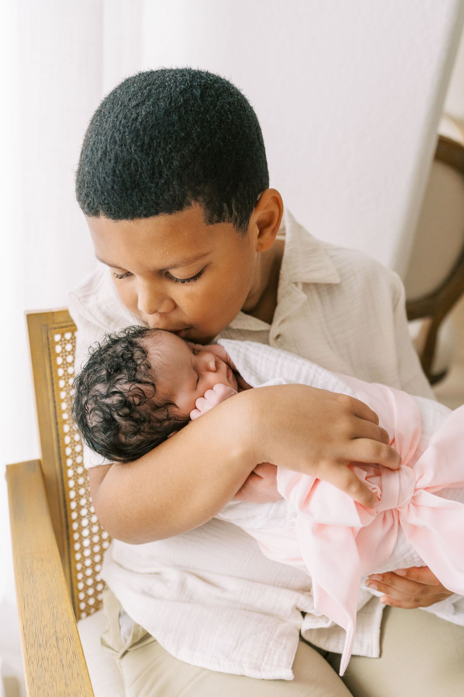 An older boy gently kisses a newborn baby wrapped in a pink blanket while seated in a chair.