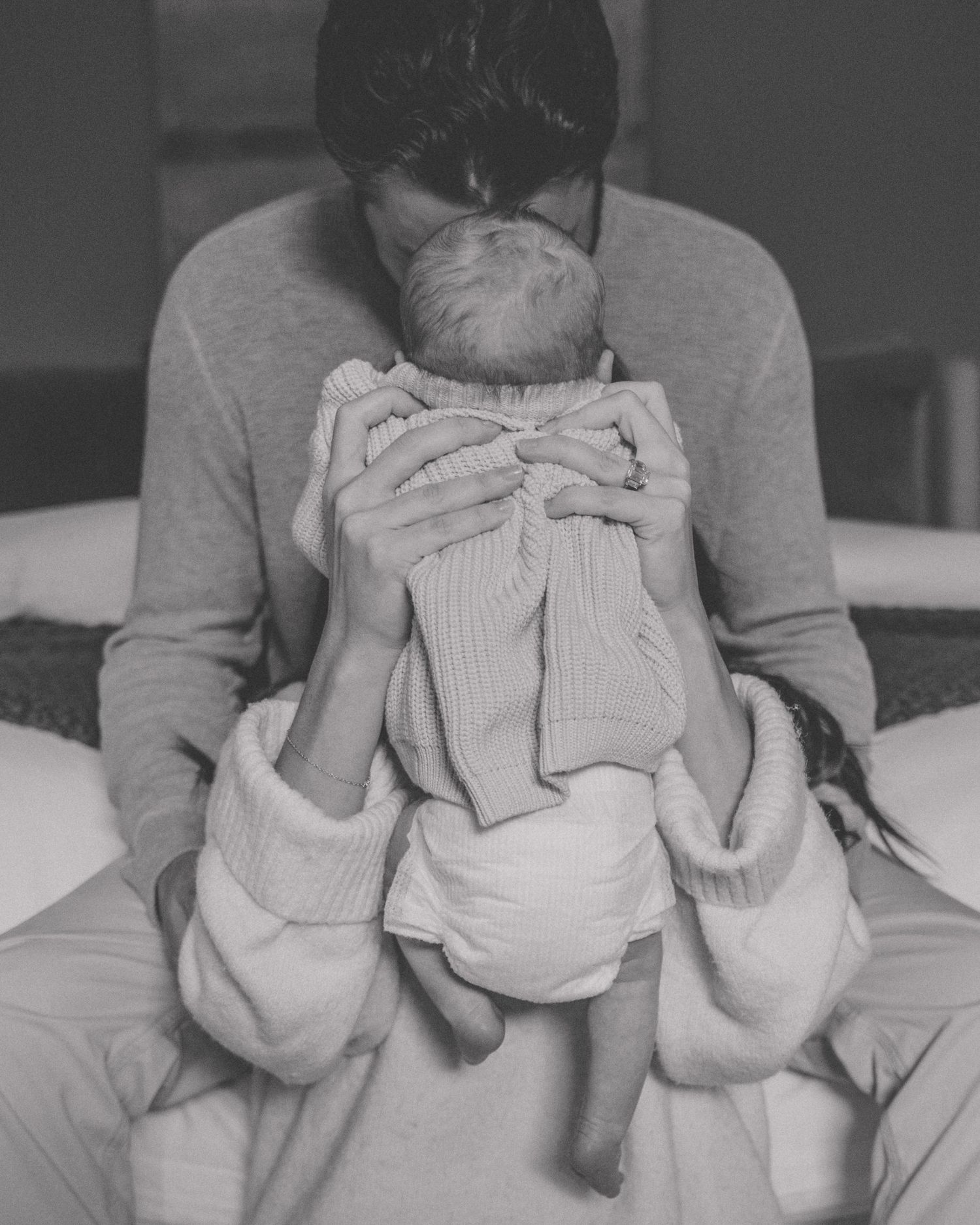 Black and white photo of a parent kissing a newborn baby wrapped in a knit blanket while sitting on a bed.