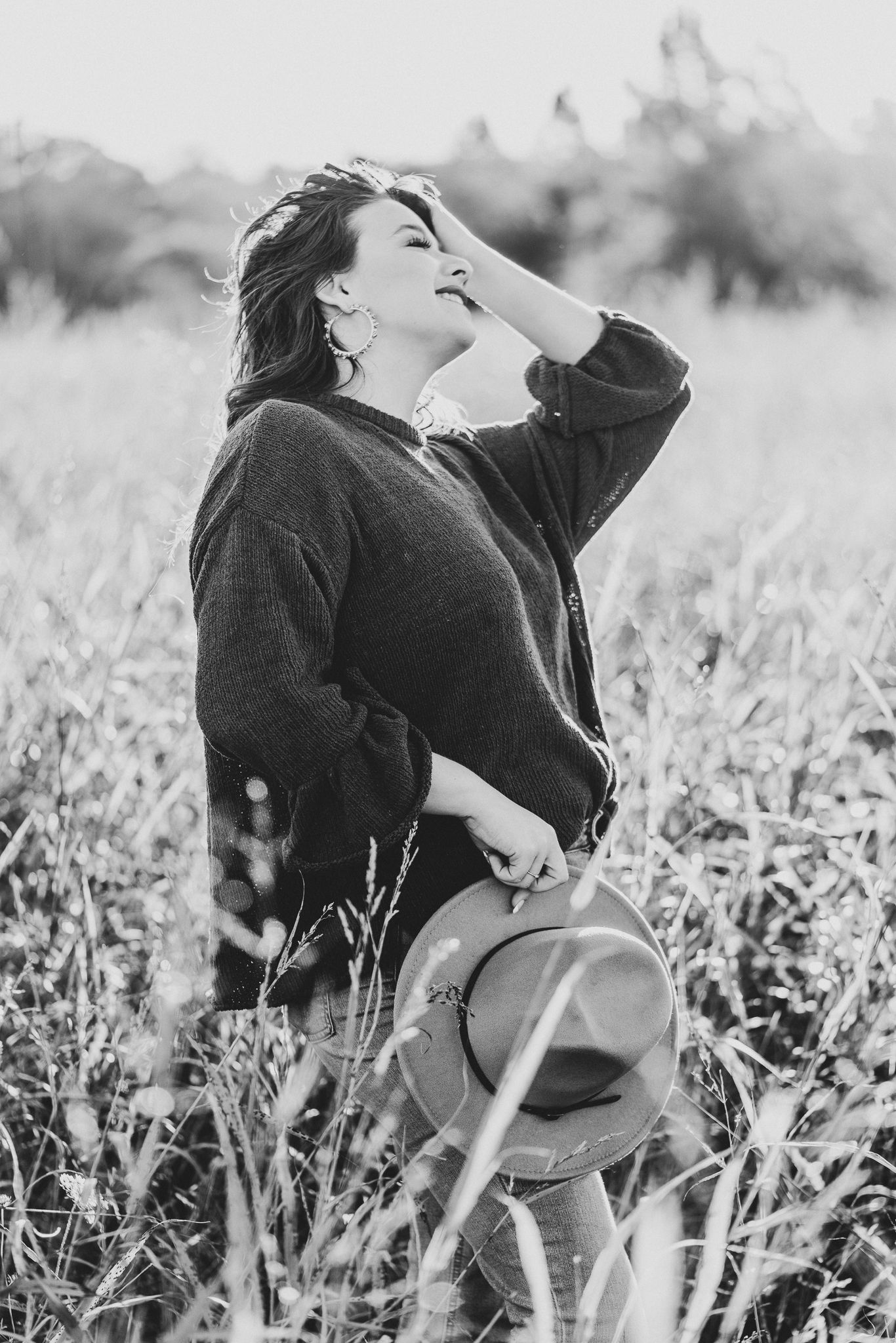 Black and white portrait of someone laughing joyfully in a field while holding a hat on a sunny day.