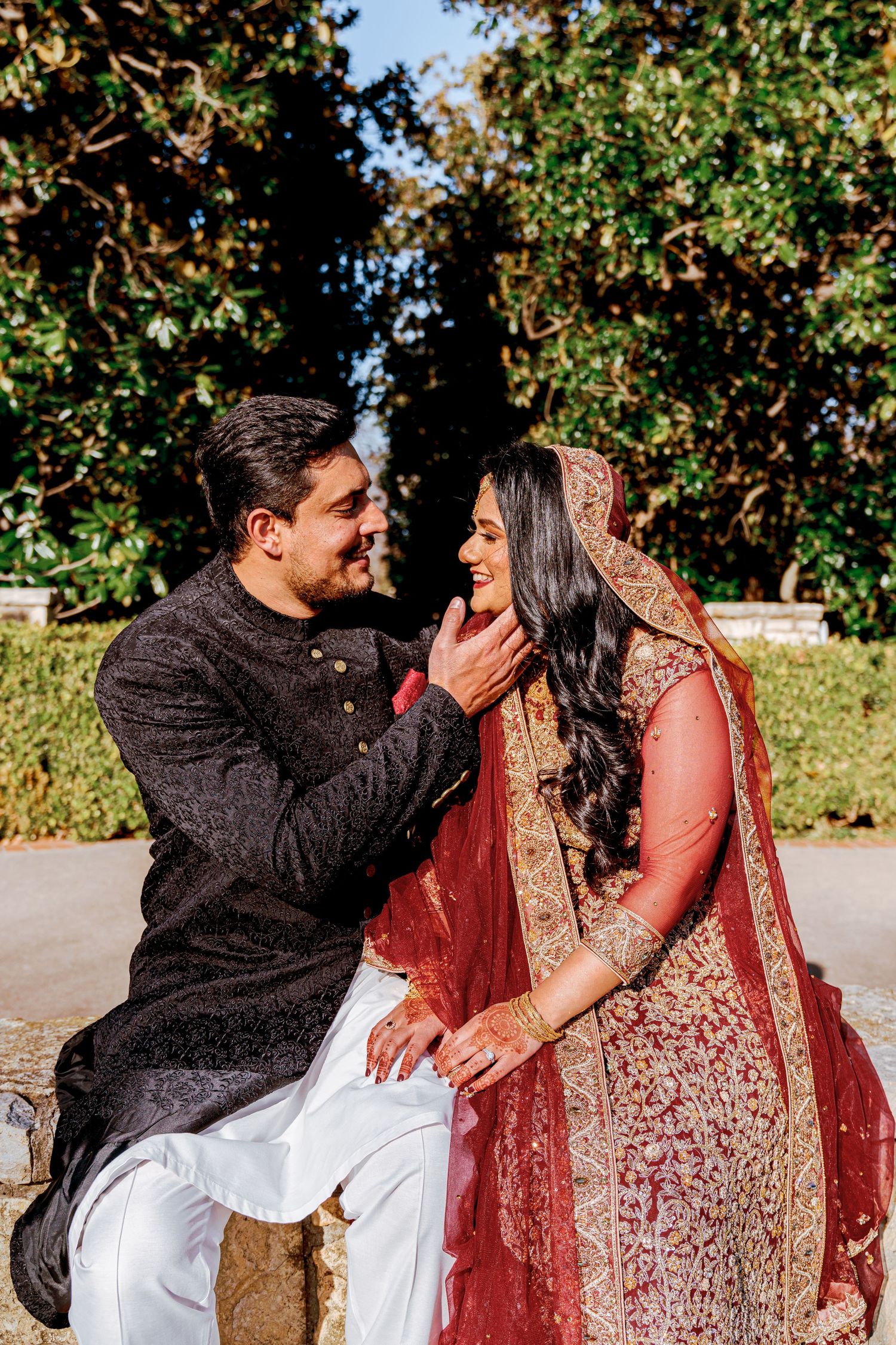 A couple in traditional South Asian attire share an intimate moment outdoors on a sunny day.