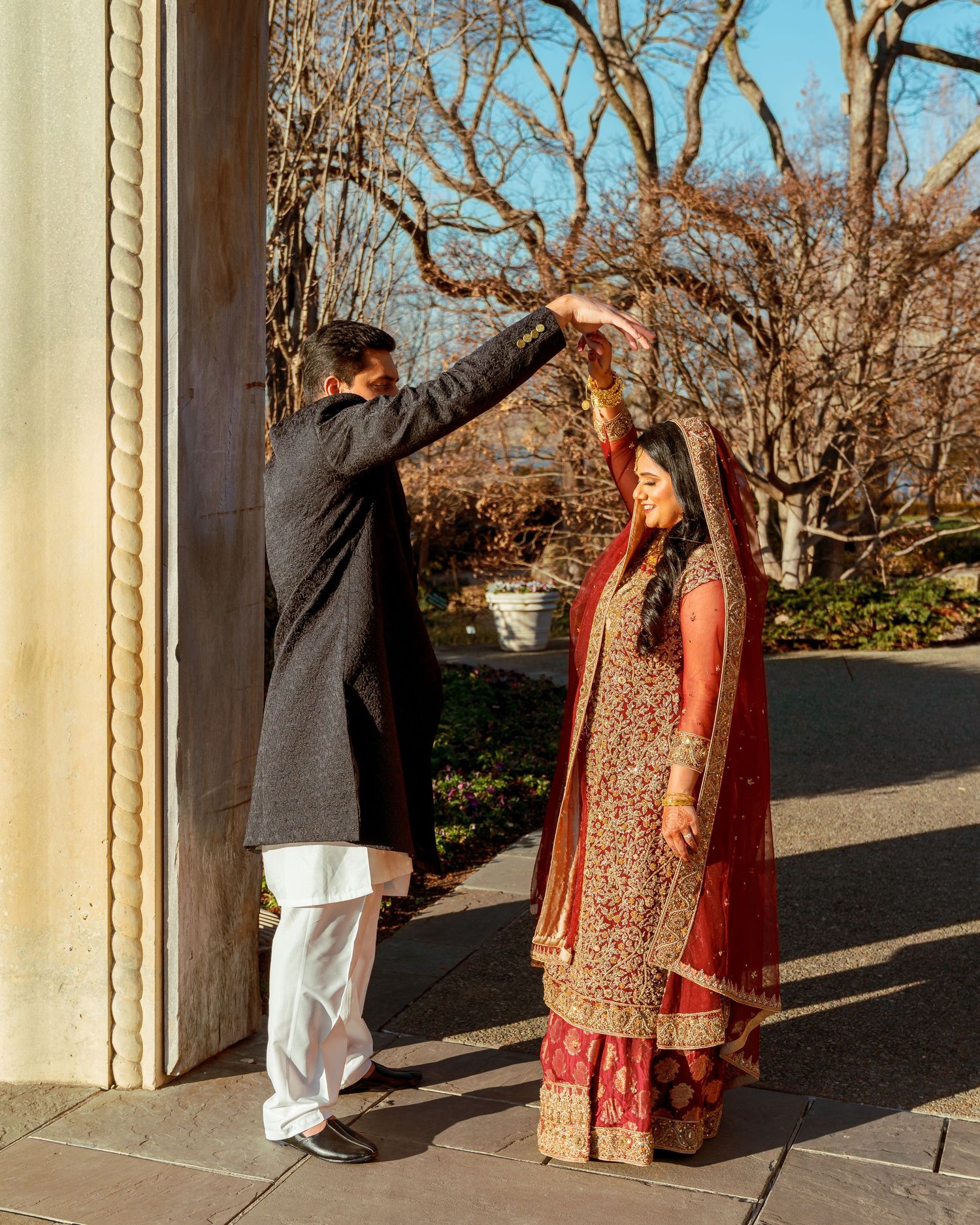 A couple in traditional South Asian wedding attire poses playfully between outdoor stone columns.
