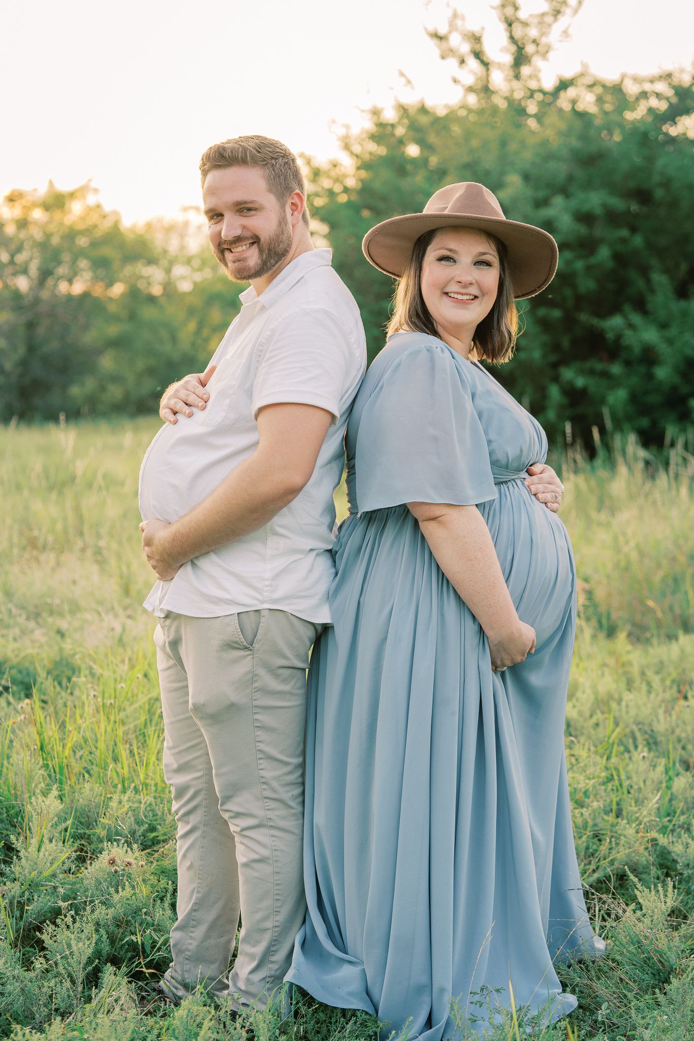 Playful maternity photo of couple standing back to back in sunny field.