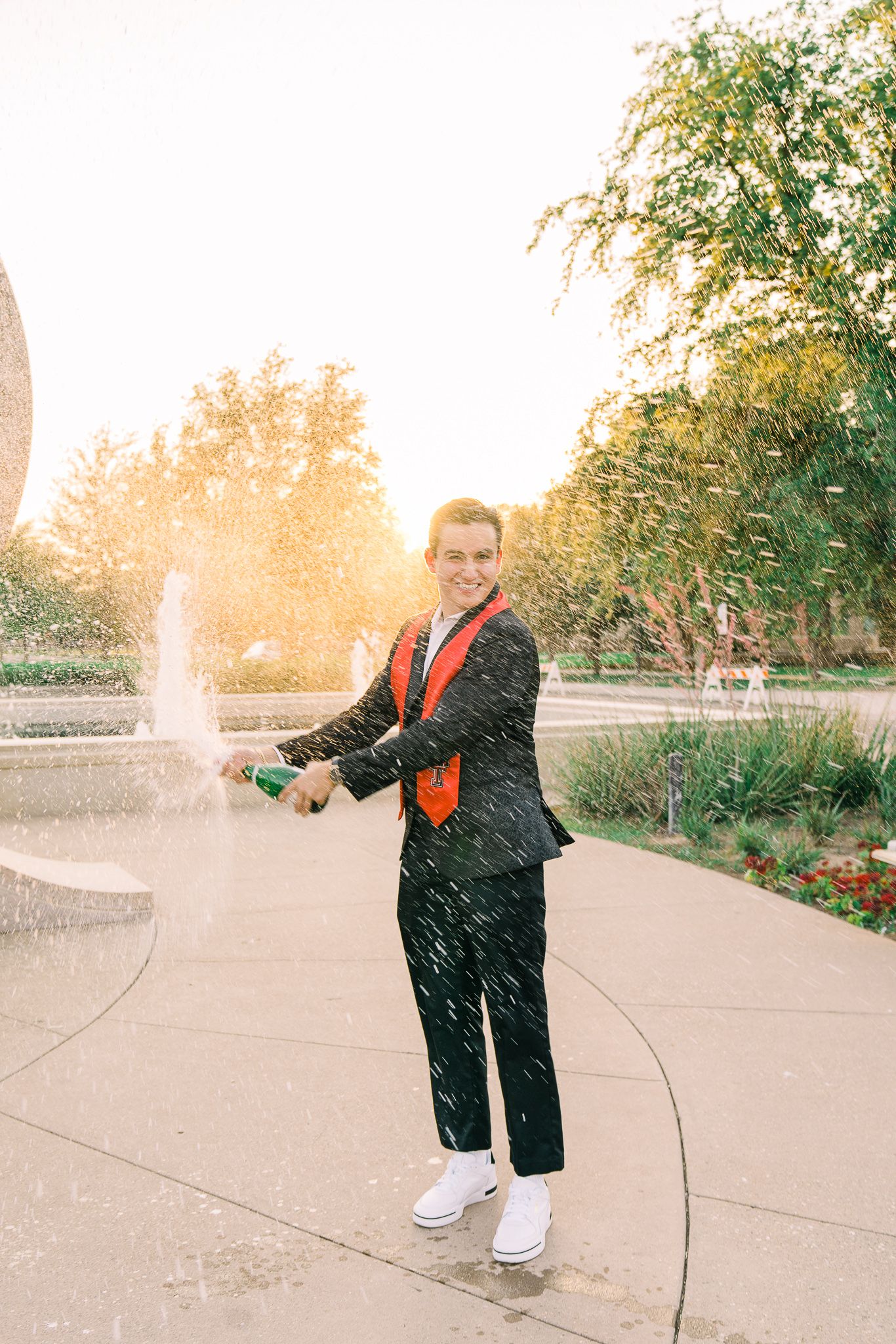 A person in a dark suit and orange tie sprays water from a fountain on a garden path at sunset.