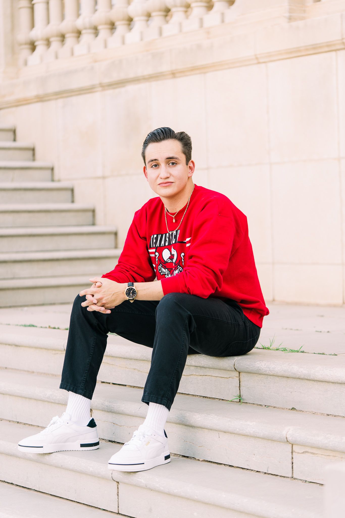 A person wearing a red sweatshirt and black pants sits casually on concrete steps while wearing white sneakers.