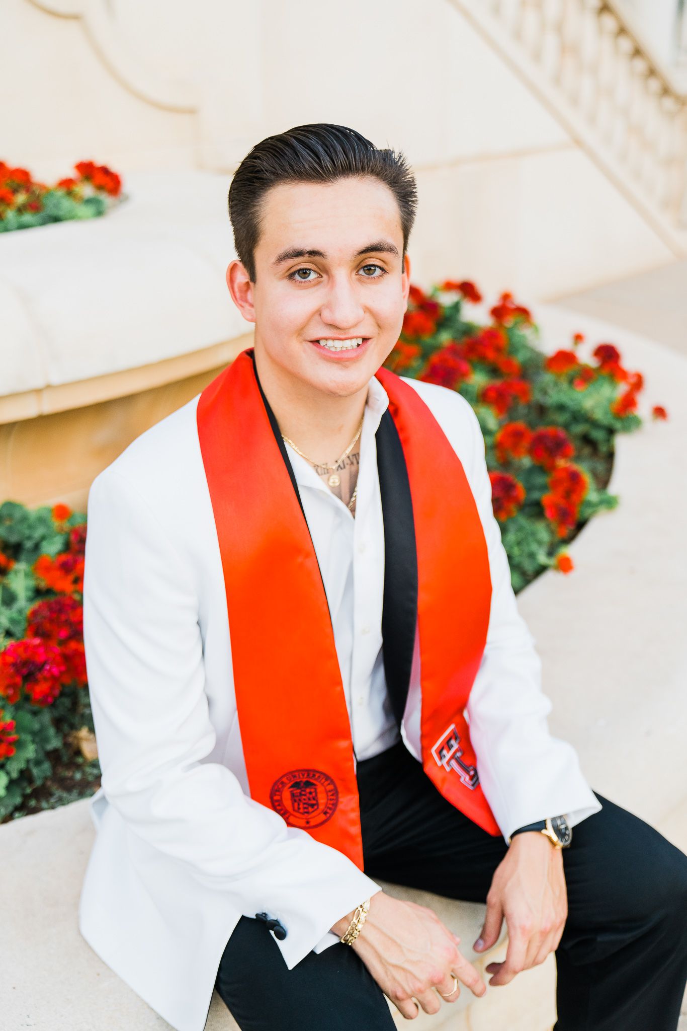 Graduate wearing red stole and white gown poses for outdoor portrait with red flowers in background.