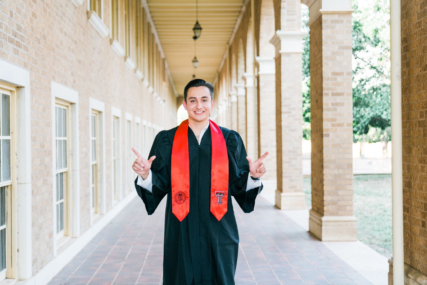 Graduate in cap and gown stands in a sunlit stone colonnade walkway with classical architecture.