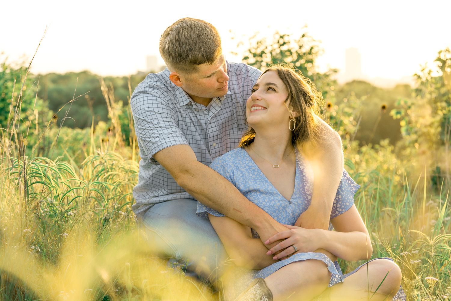 A romantic couple embraces in a sunlit meadow during golden hour while surrounded by wildflowers and tall grass.