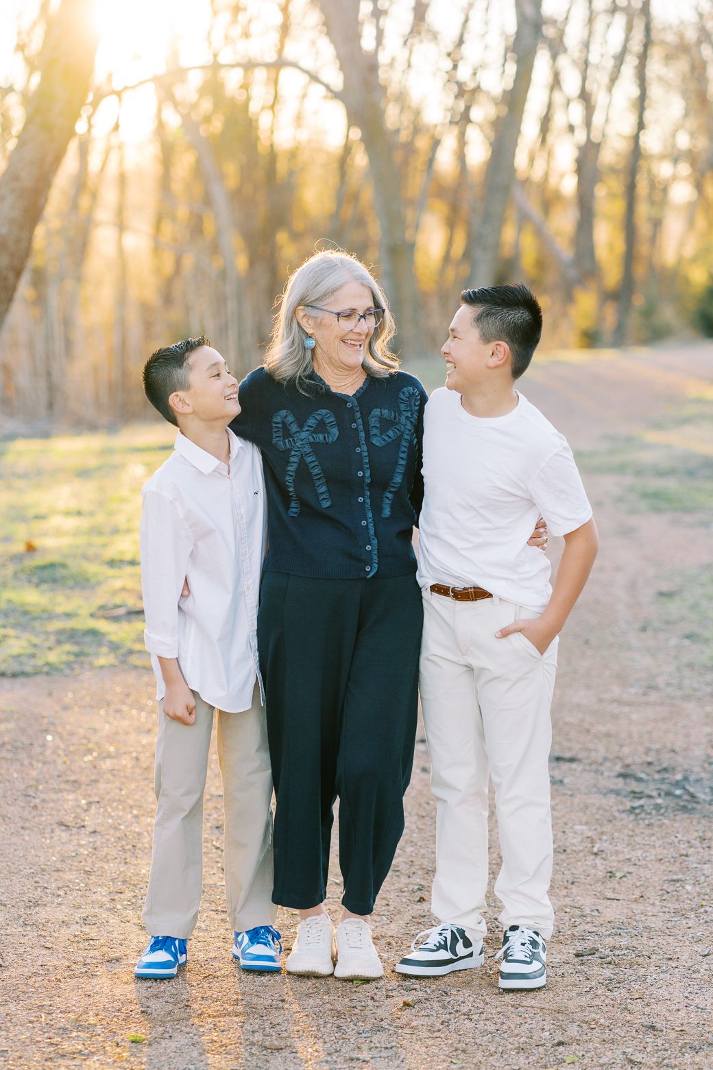 Three pairs of legs wearing casual outfits stand together on a dirt path during golden hour in a wooded setting.