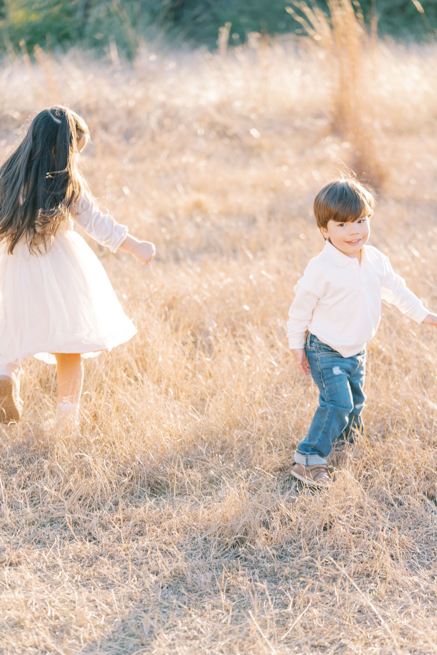 A child in a white top and jeans runs playfully through tall golden grass at sunset.