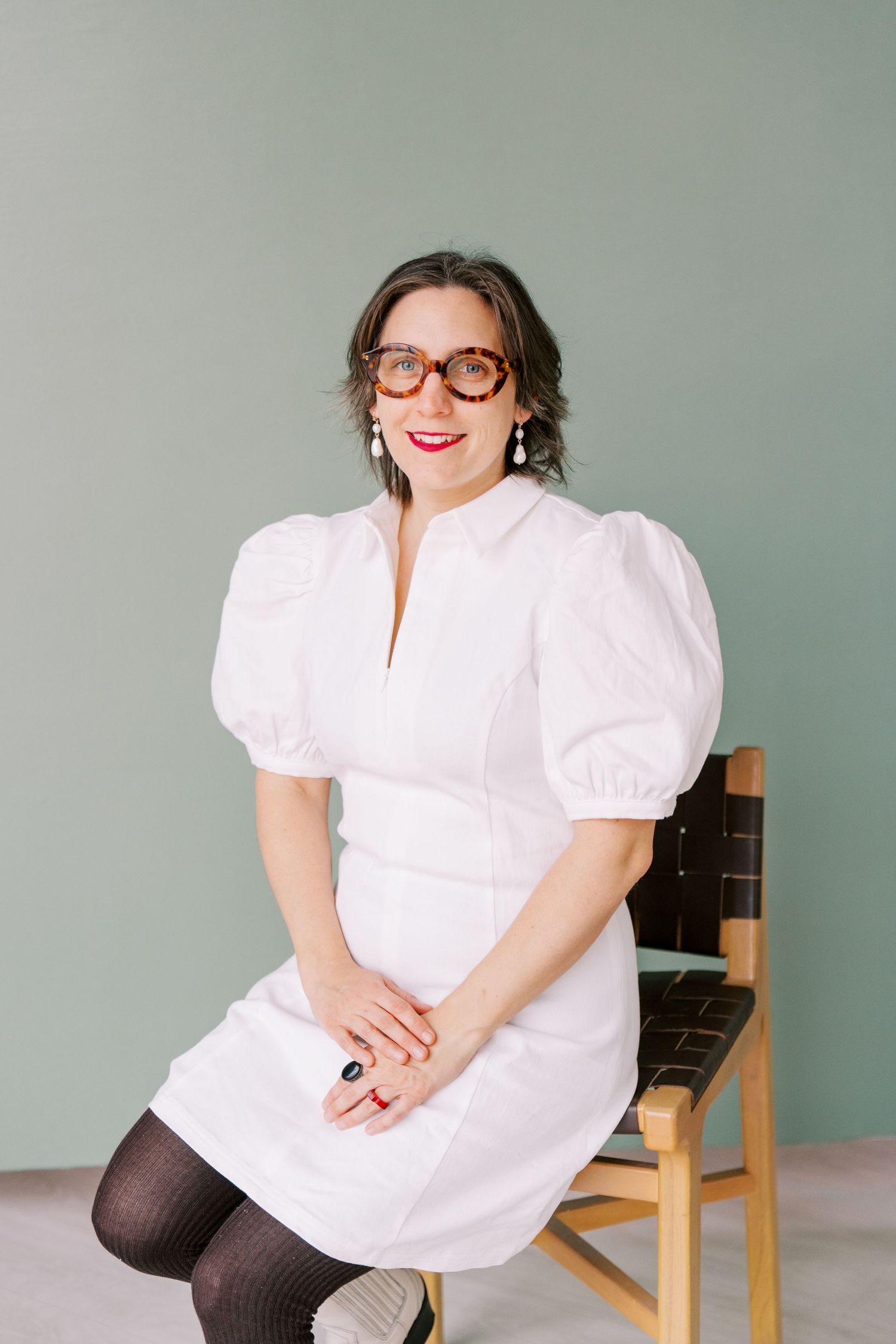 A person in a crisp white blouse and glasses sits on a wooden chair against a grey backdrop.