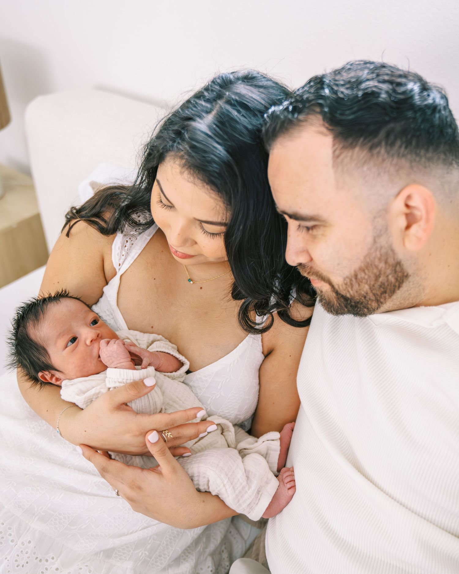 New parents lovingly gaze at their newborn baby wrapped in white, sharing a tender family moment on a bed.