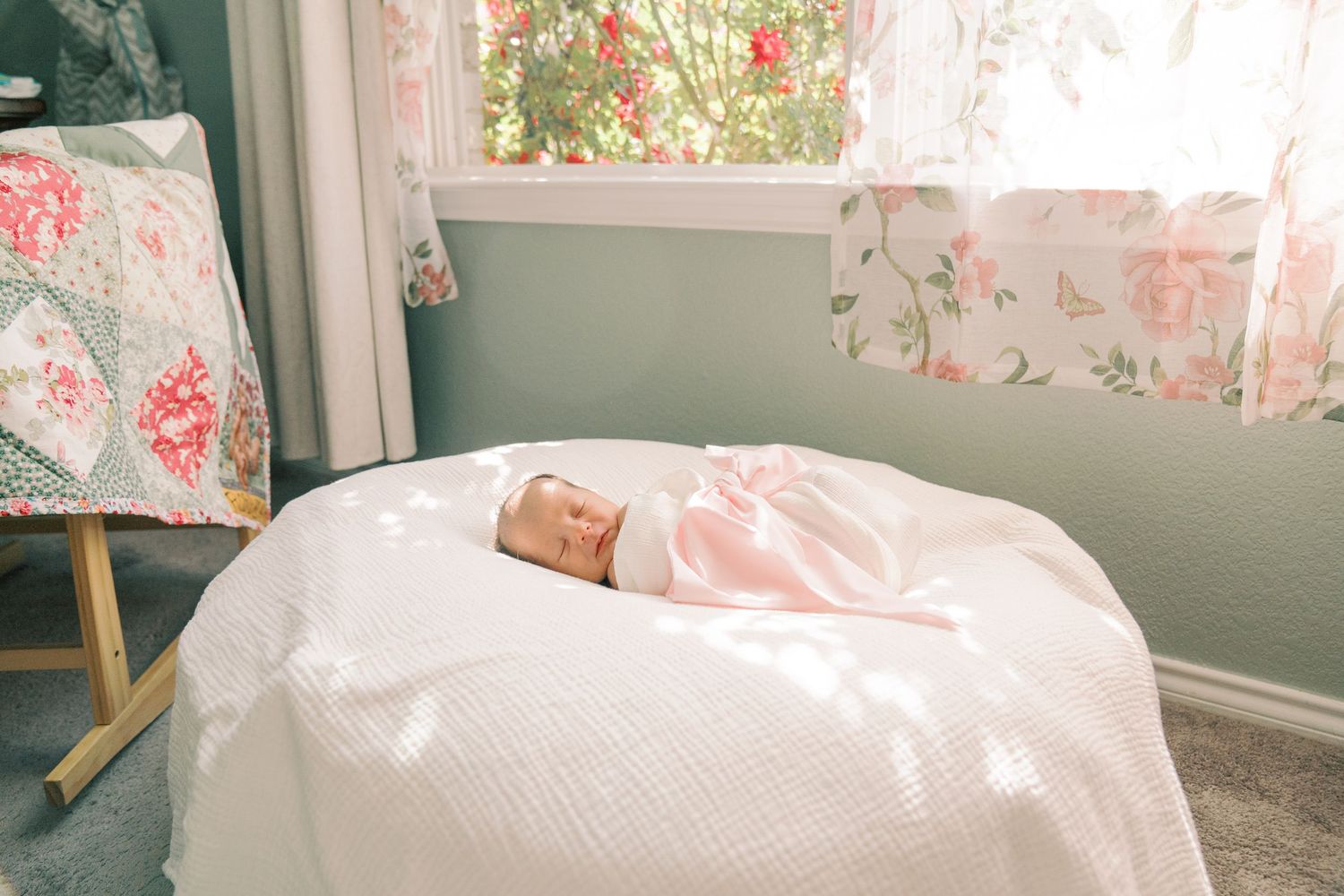 Newborn baby swaddled in pink sleeping peacefully on a white bed near a window with floral curtains.