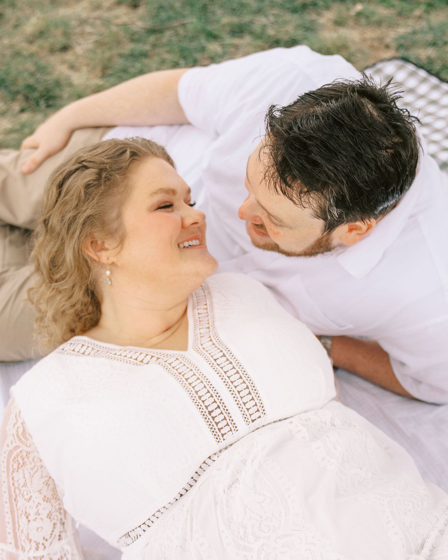 Couple lying on a blanket outdoors, woman in white lace dress smiling up at man in white shirt lovingly.