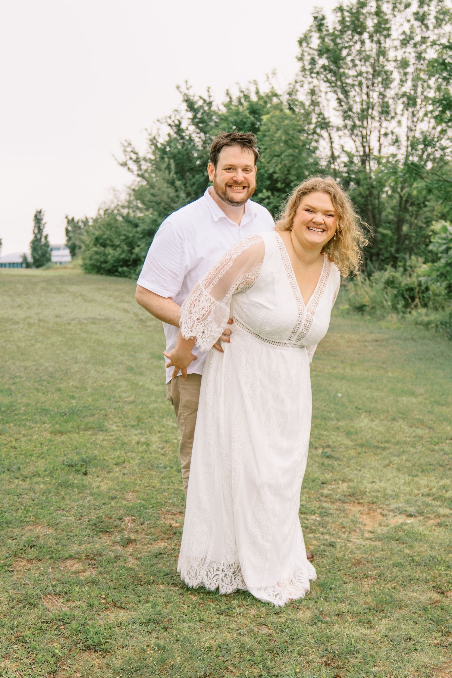 Happy couple posing together on green lawn, woman in white lace dress and man in white button-up shirt.