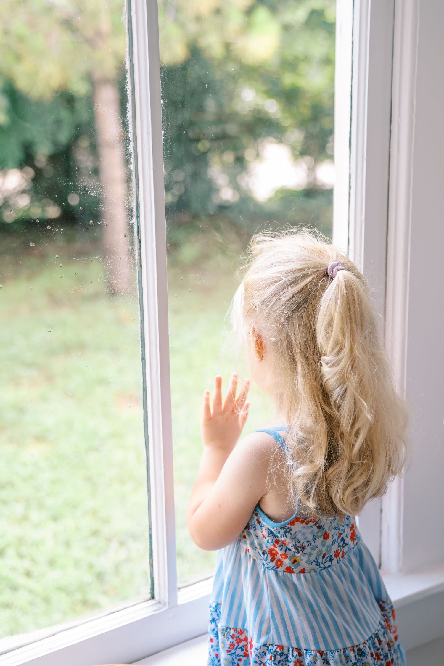 Young blonde girl in floral dress looking out a window at green yard, hand pressed against glass.