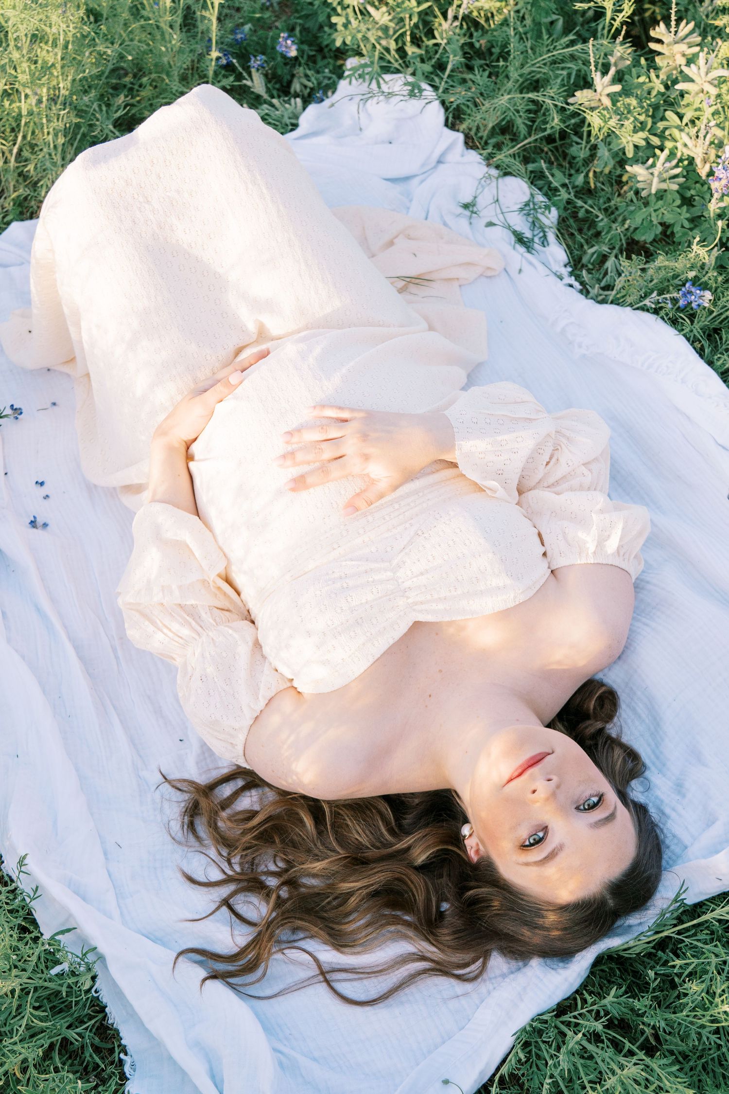 Pregnant woman in white dress lying on blanket in green field, cradling baby bump with flowing hair spread around her.