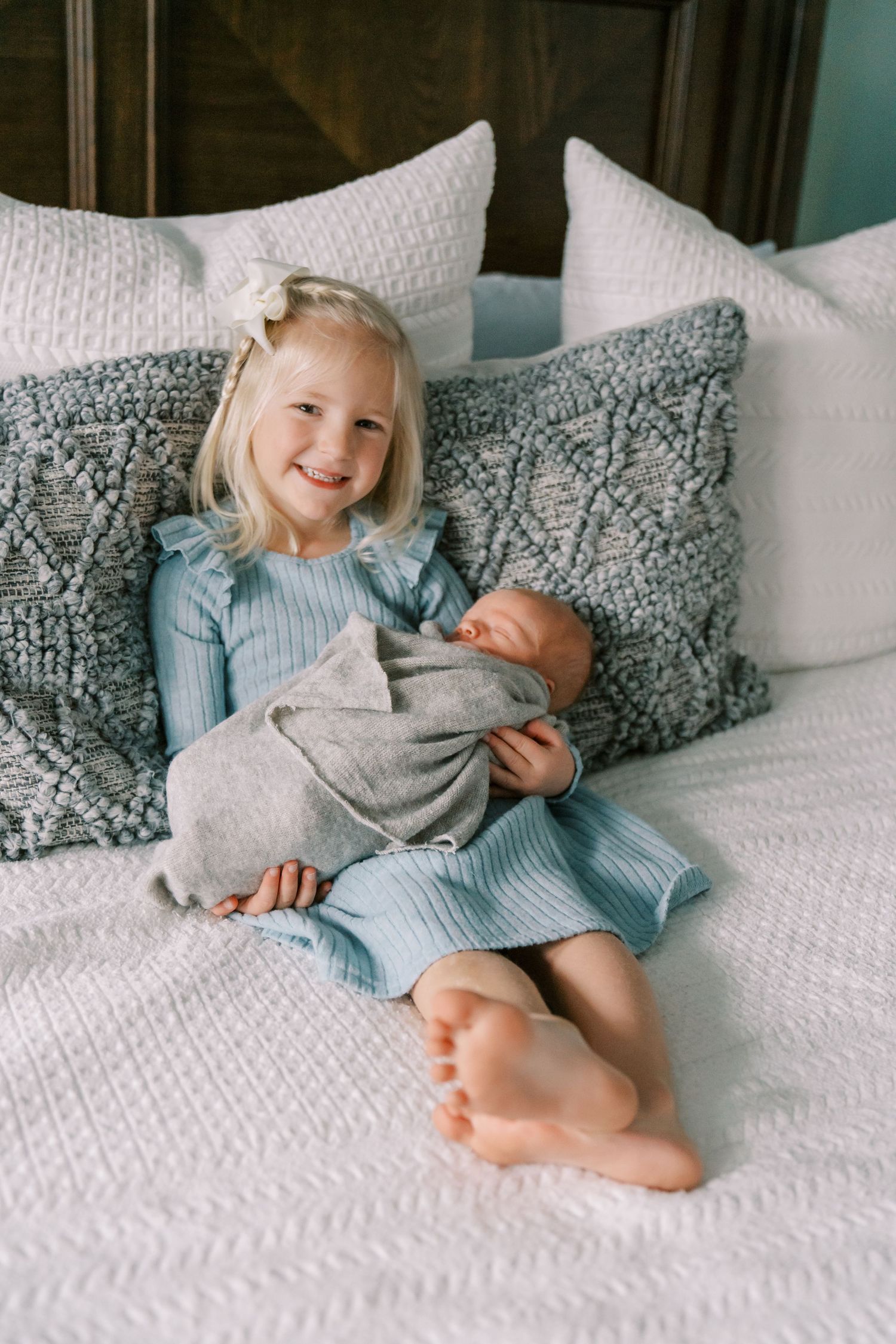 Young child in blue dress sits on white bedding while holding a small baby wrapped in gray blanket.