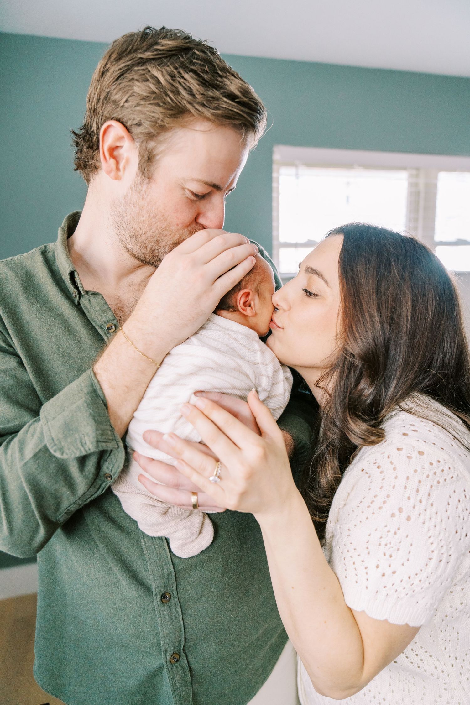 Parents lovingly kiss their newborn baby while standing together in a room with turquoise walls.