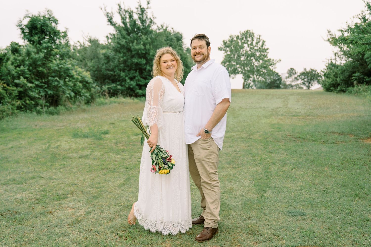 Smiling couple posing in green field, woman holding colorful bouquet in white lace dress outdoors.