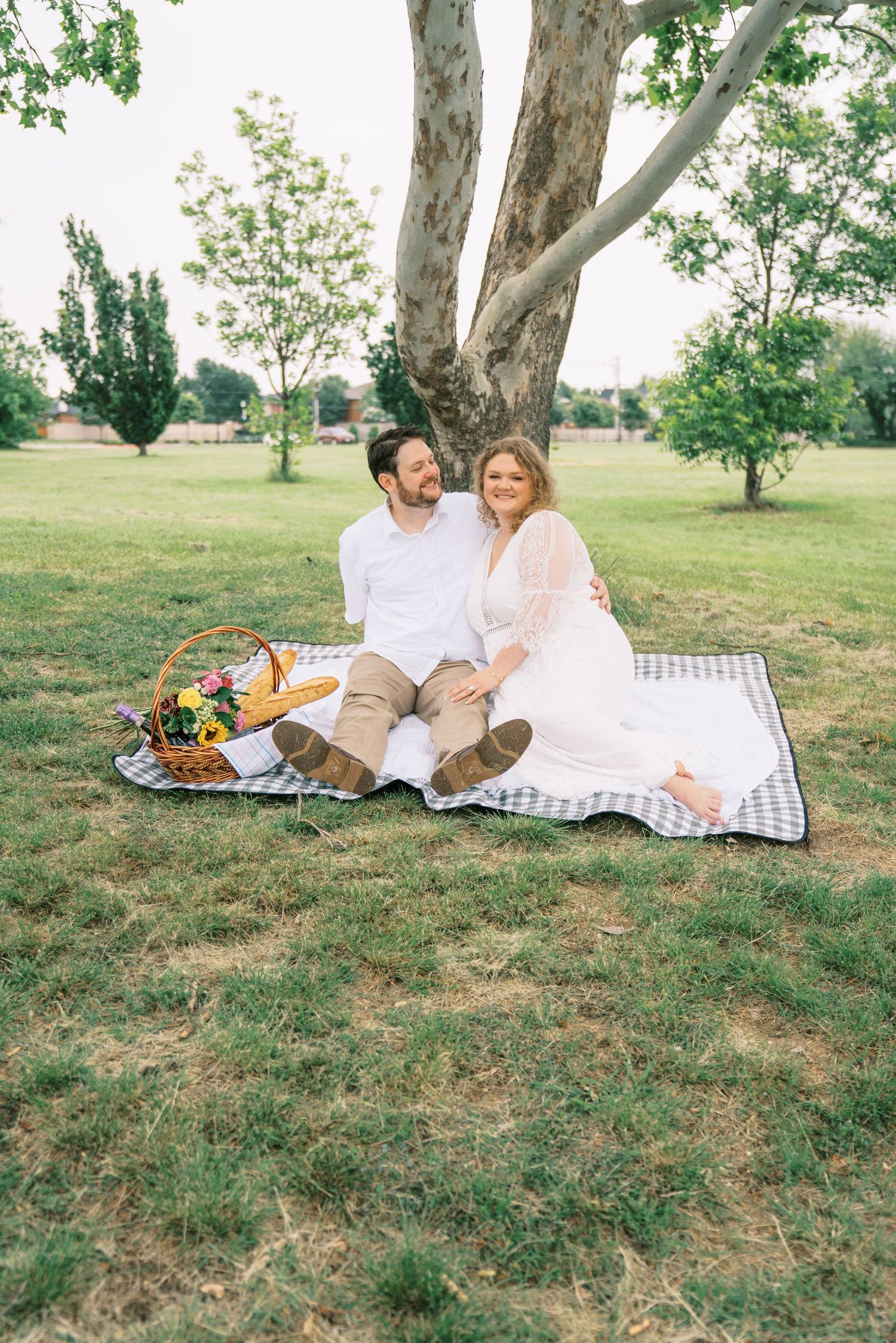 Couple sitting on a checkered blanket under a tree during a romantic outdoor picnic with a wicker basket nearby.