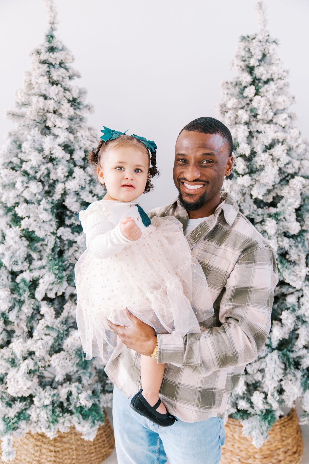 Smiling father holding young daughter in white dress before snow-flocked Christmas trees in bright studio setting.