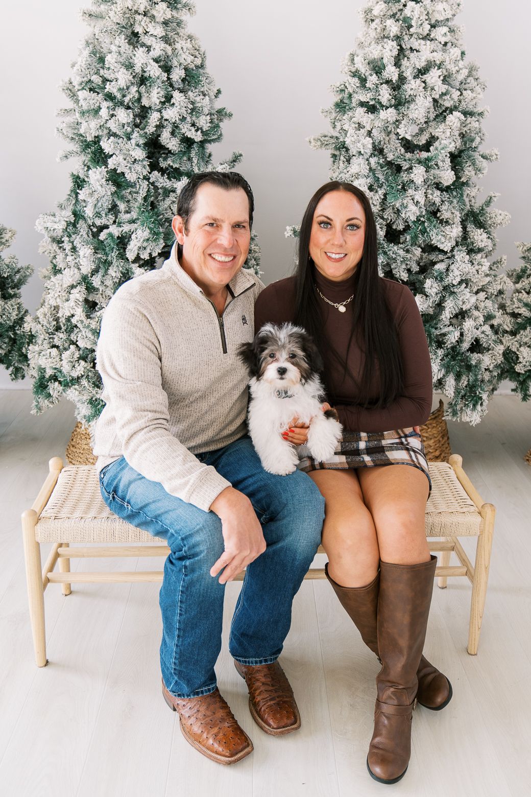 A couple sits on a wooden bench with their small white dog in front of a snow-flocked Christmas tree for holiday photos.