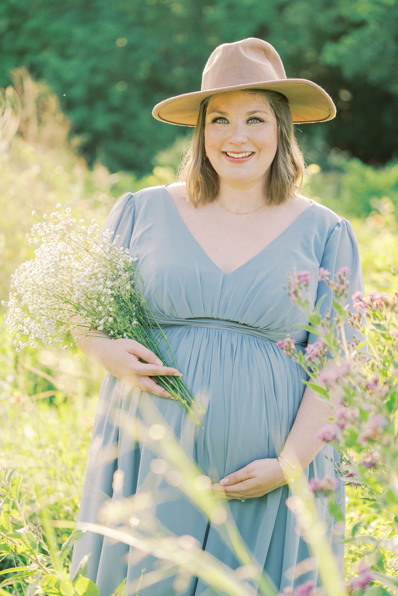 A person wearing a light blue empire waist dress and sun hat poses among blooming wildflowers in a meadow.