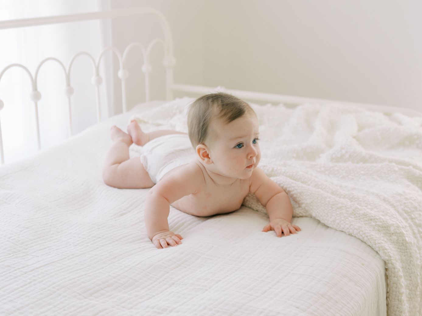 Baby in white onesie crawls on soft white bedding while exploring the cozy space.