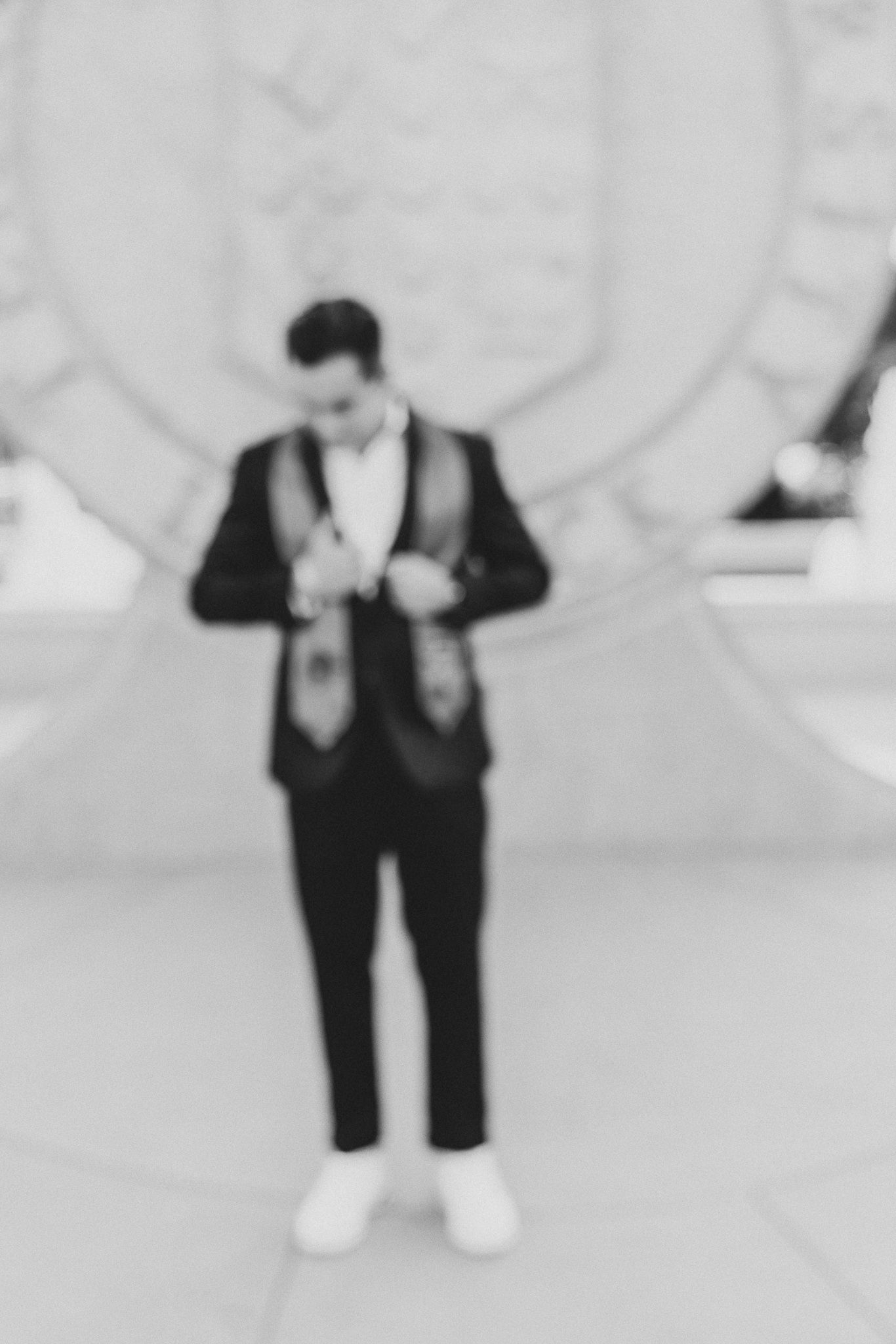 Blurry black and white photo of someone in a formal suit and white sneakers standing against a light background.