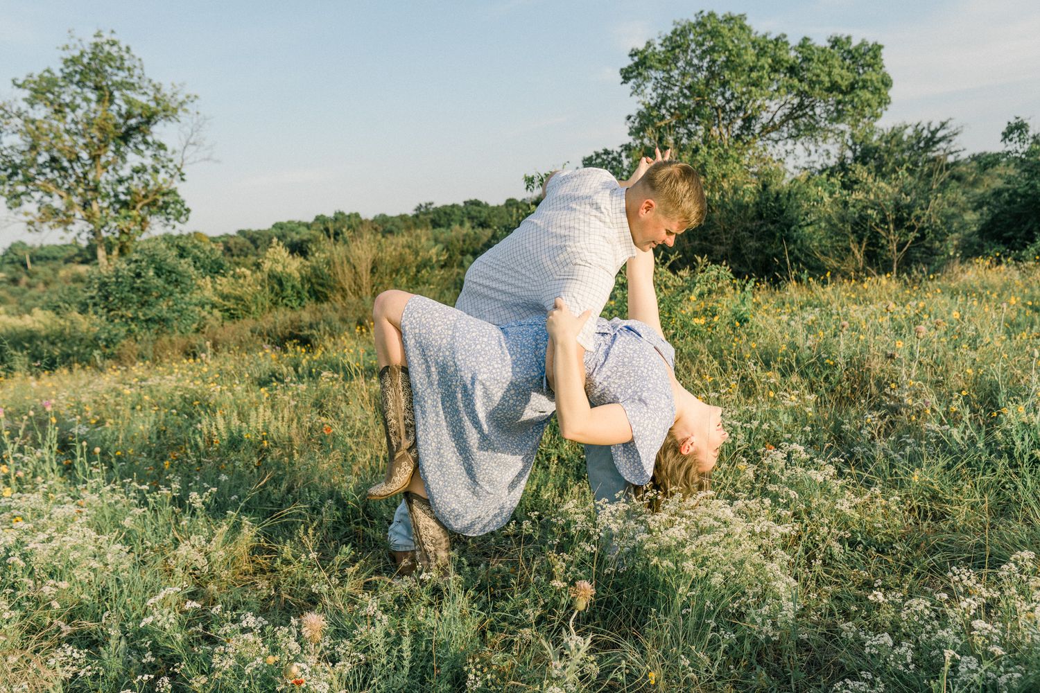 Two people hold hands while running through a green summer field with trees in the background.