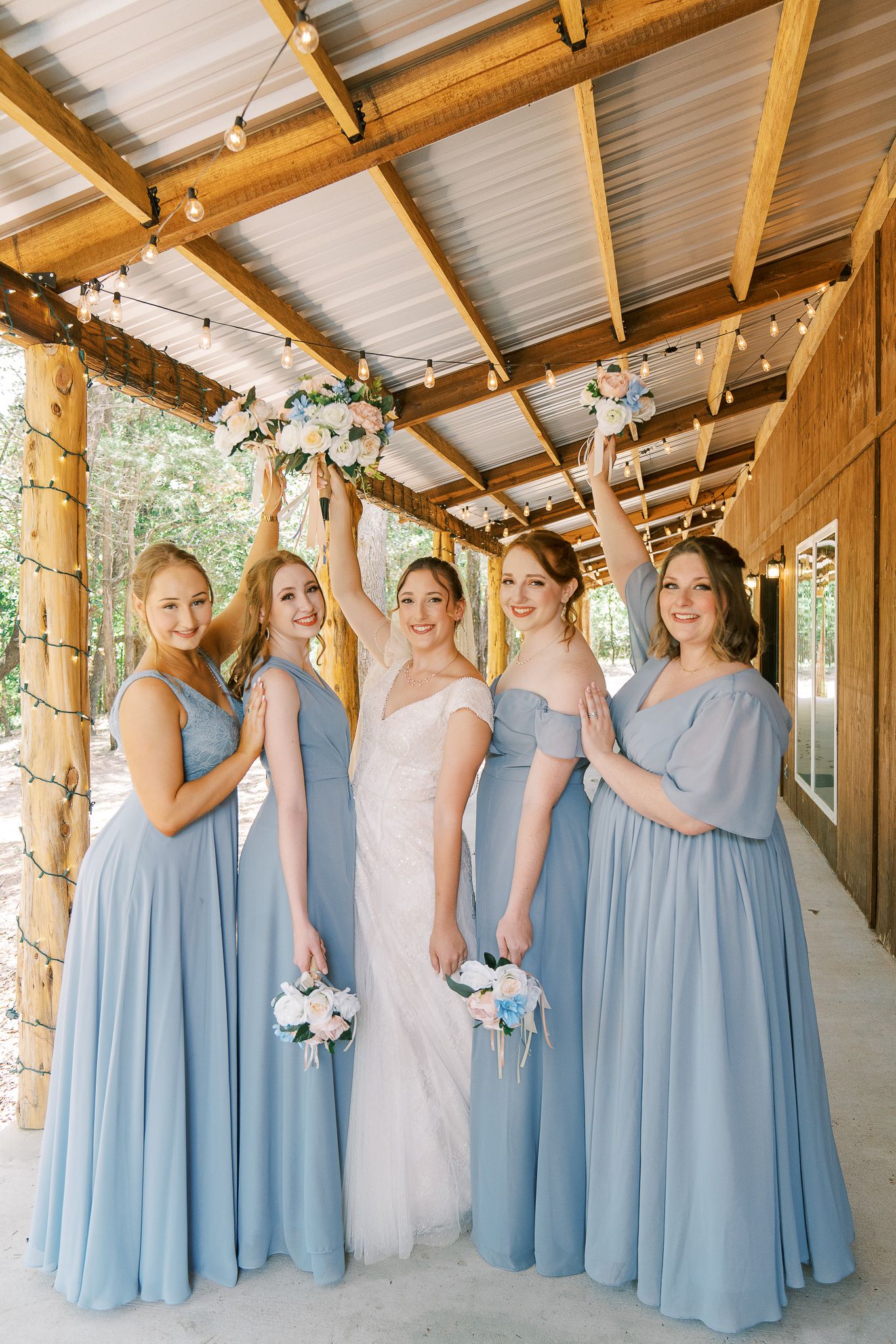 Wedding party wearing light blue bridesmaid dresses and white gown pose together in rustic wooden venue.
