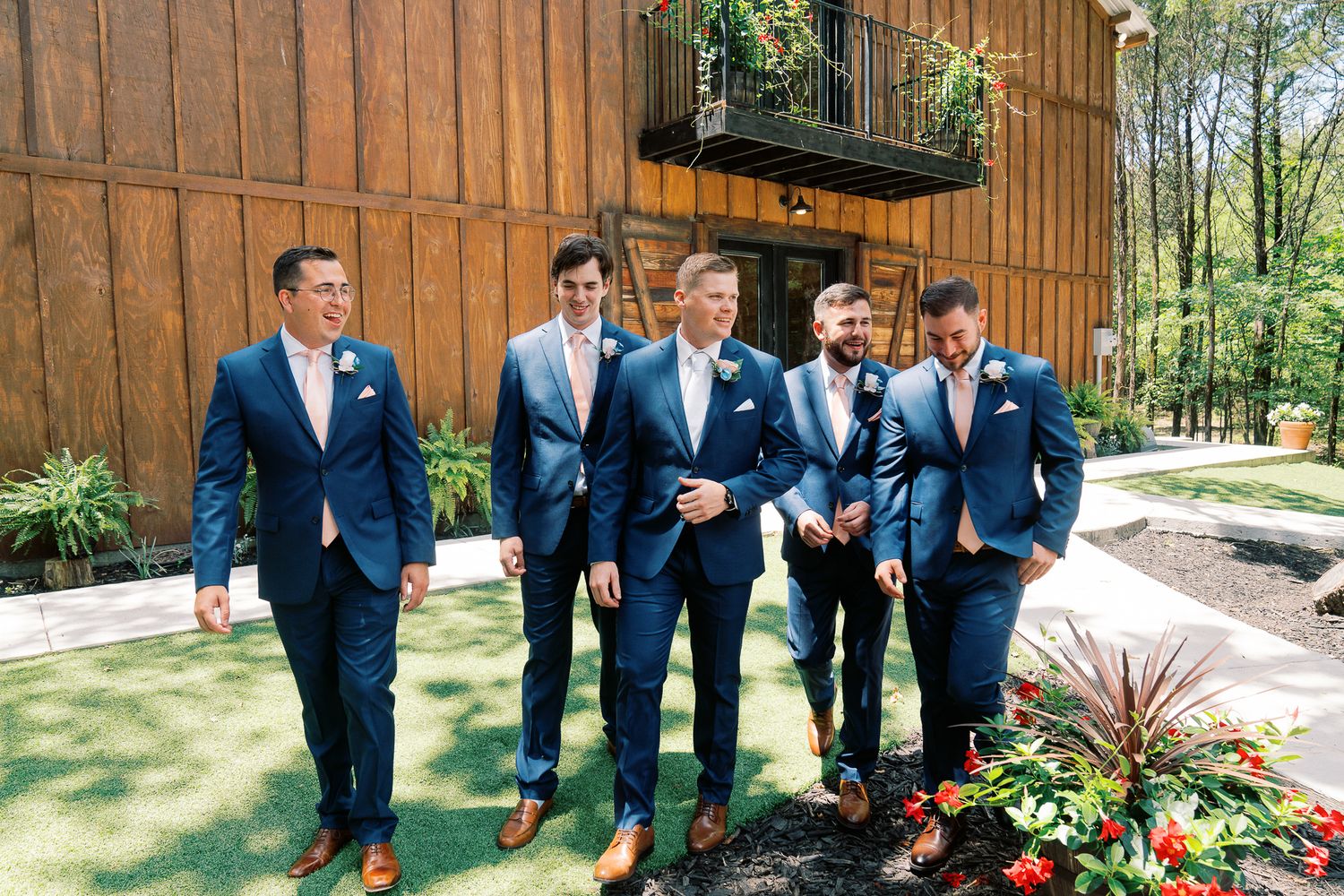Groomsmen in matching navy blue suits walk together and laugh on a sunny day near a rustic barn venue.