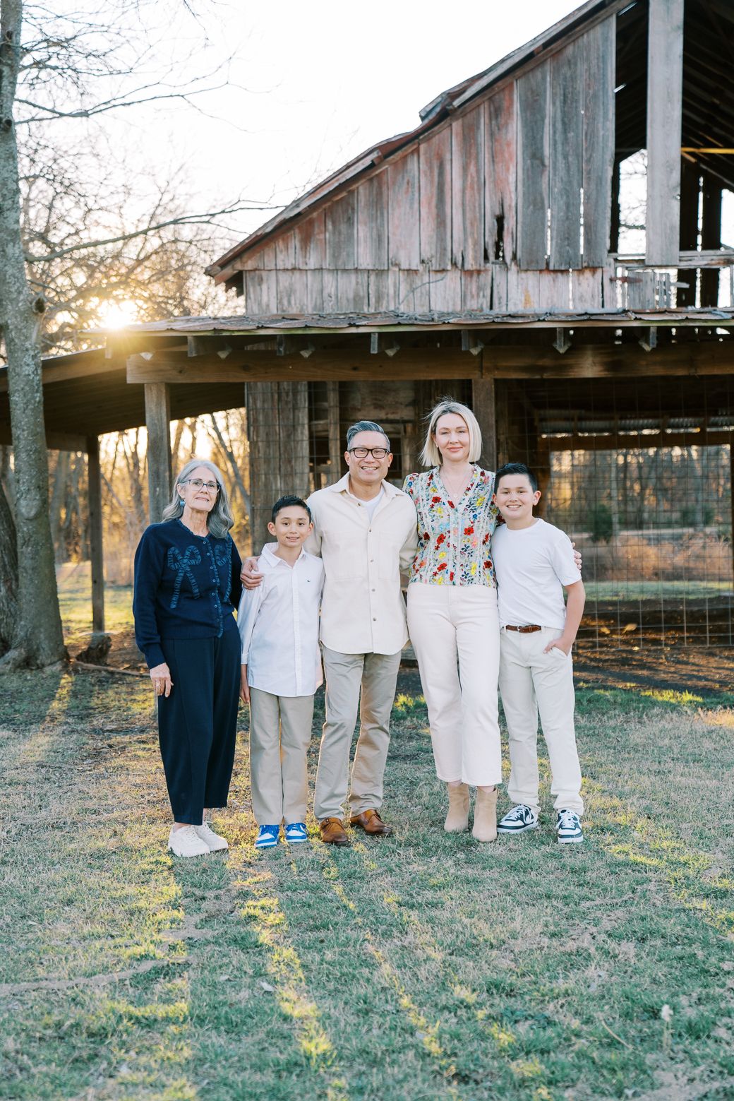 A group stands together outdoors in coordinated neutral outfits against a rustic wooden barn backdrop at sunset.
