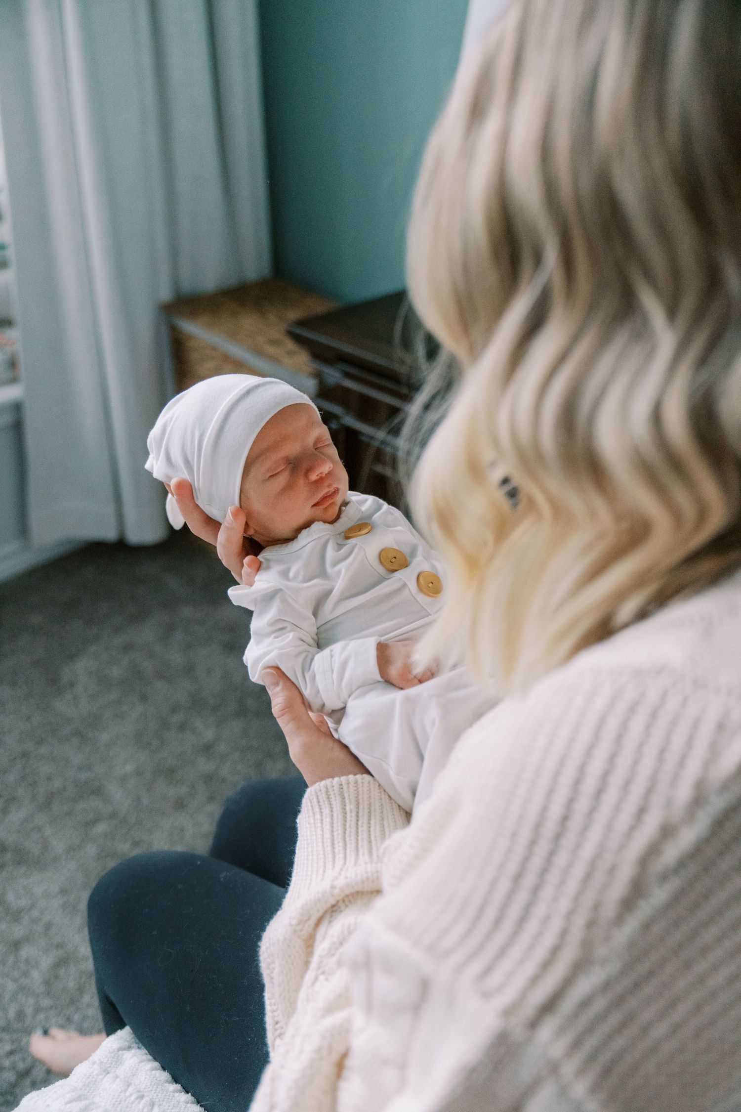 Mother in cream sweater cradling newborn baby wearing white outfit and bonnet in nursery.