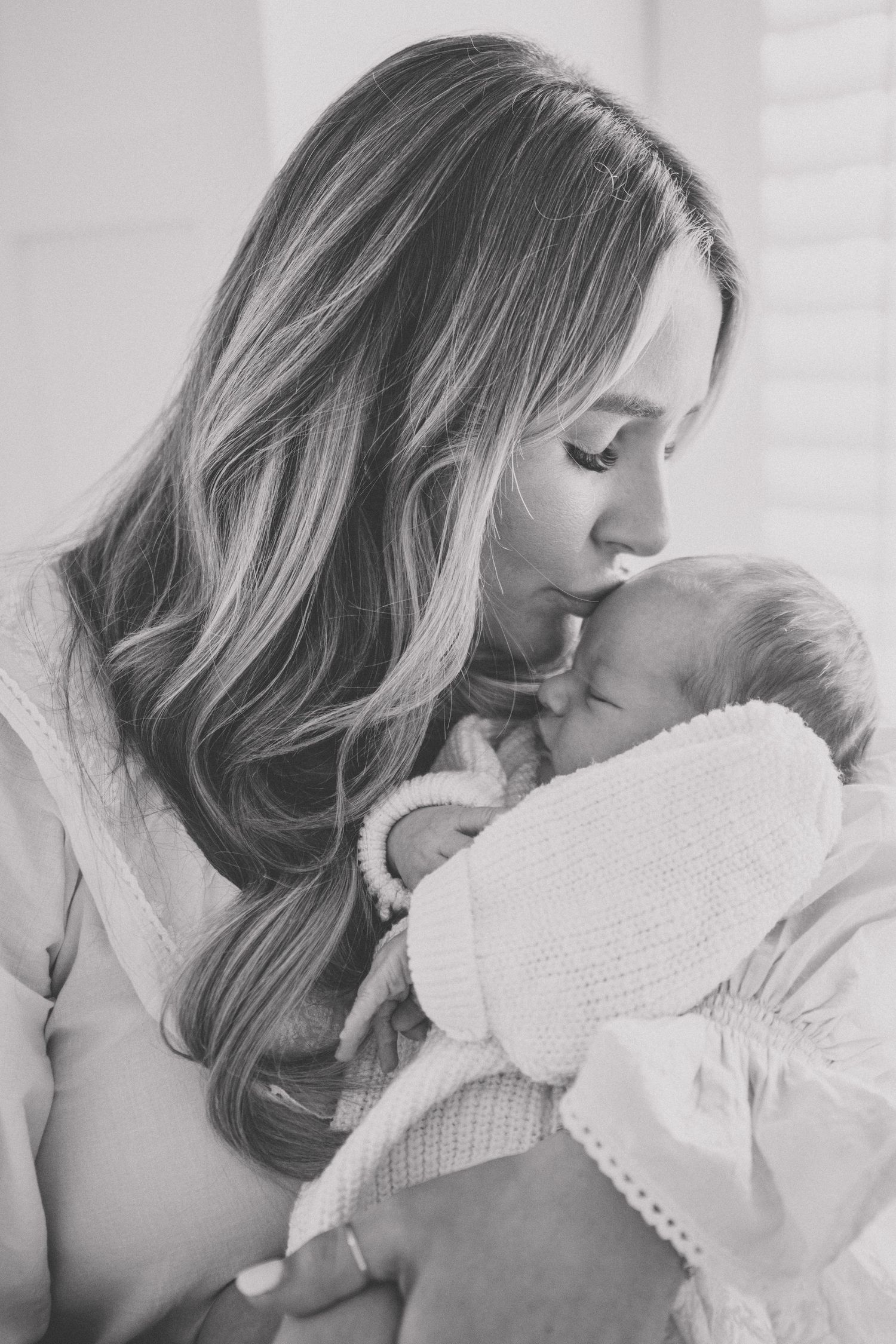 Black and white portrait of mother kissing newborn baby wrapped in blanket.