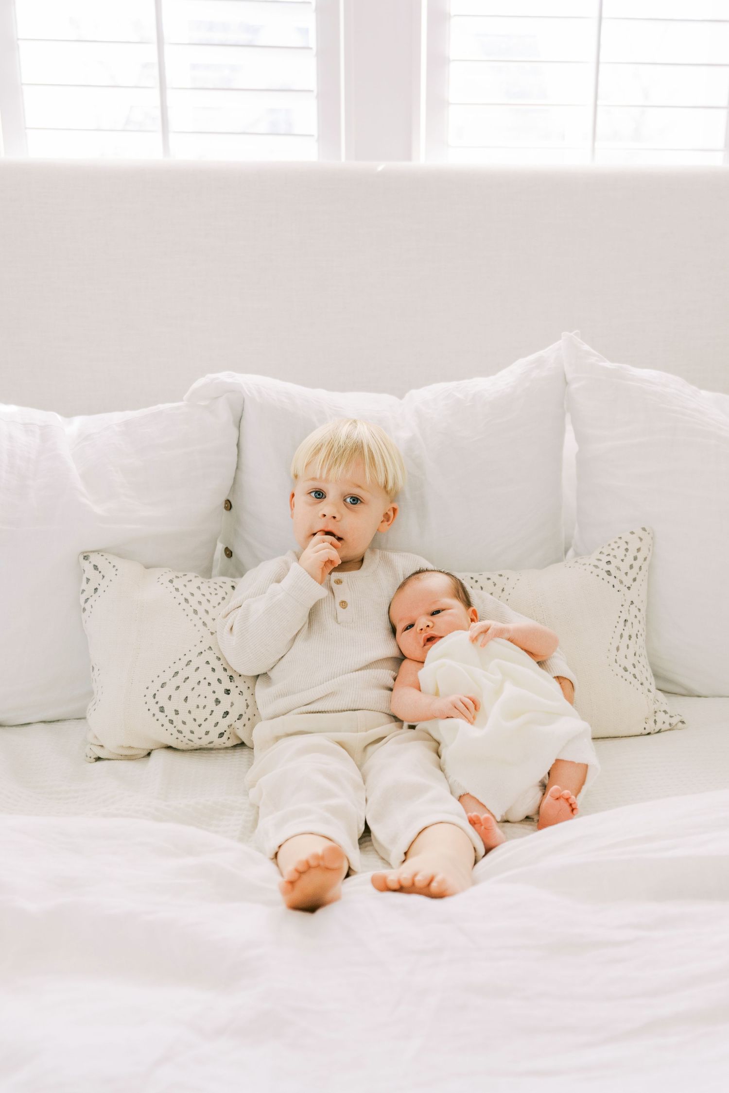 Baby in white clothing rolls and plays on a bright white bed with decorative pillows.