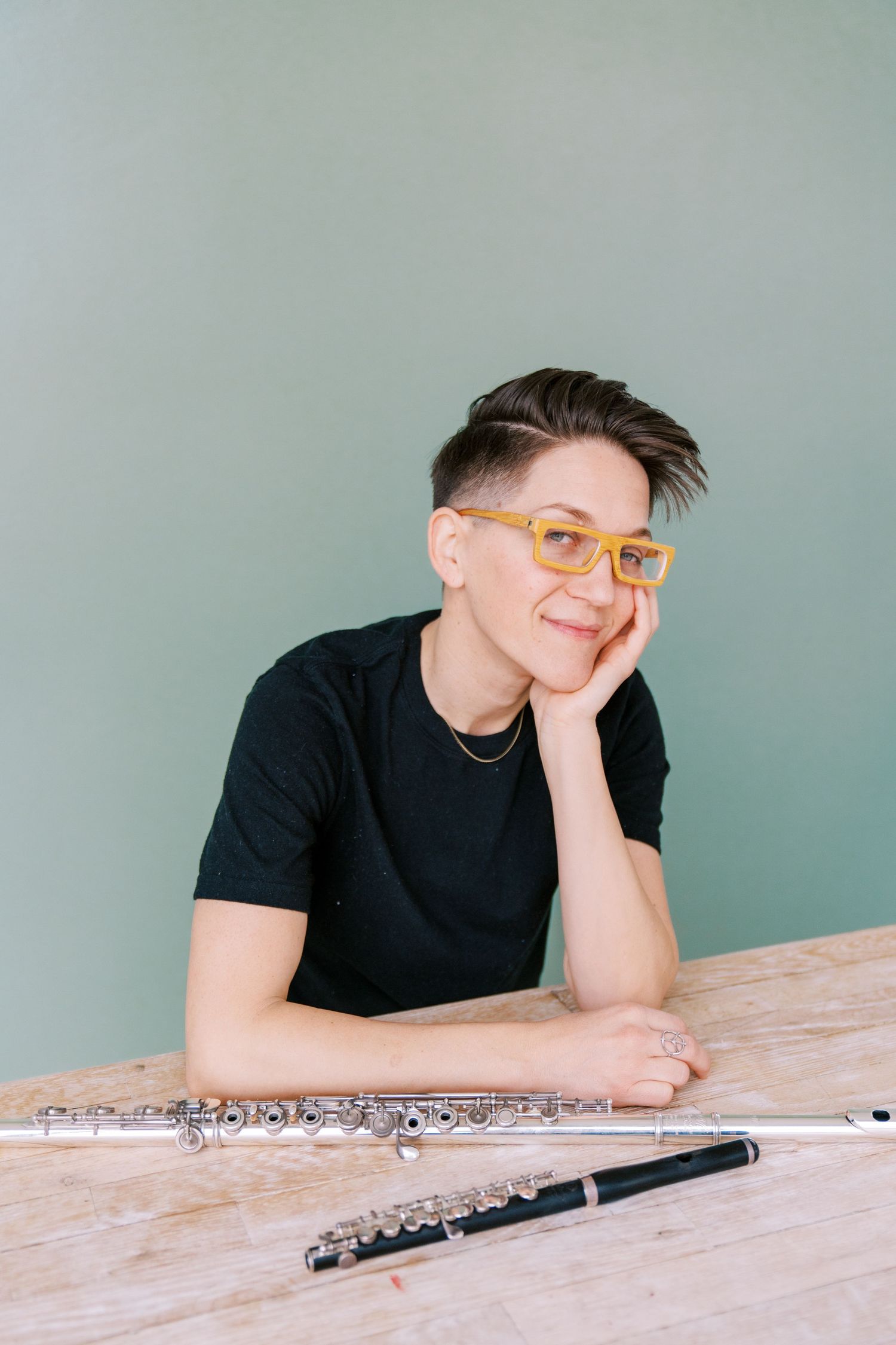 Musician in black t-shirt rests at wooden table with flute, wearing yellow glasses against mint green wall.