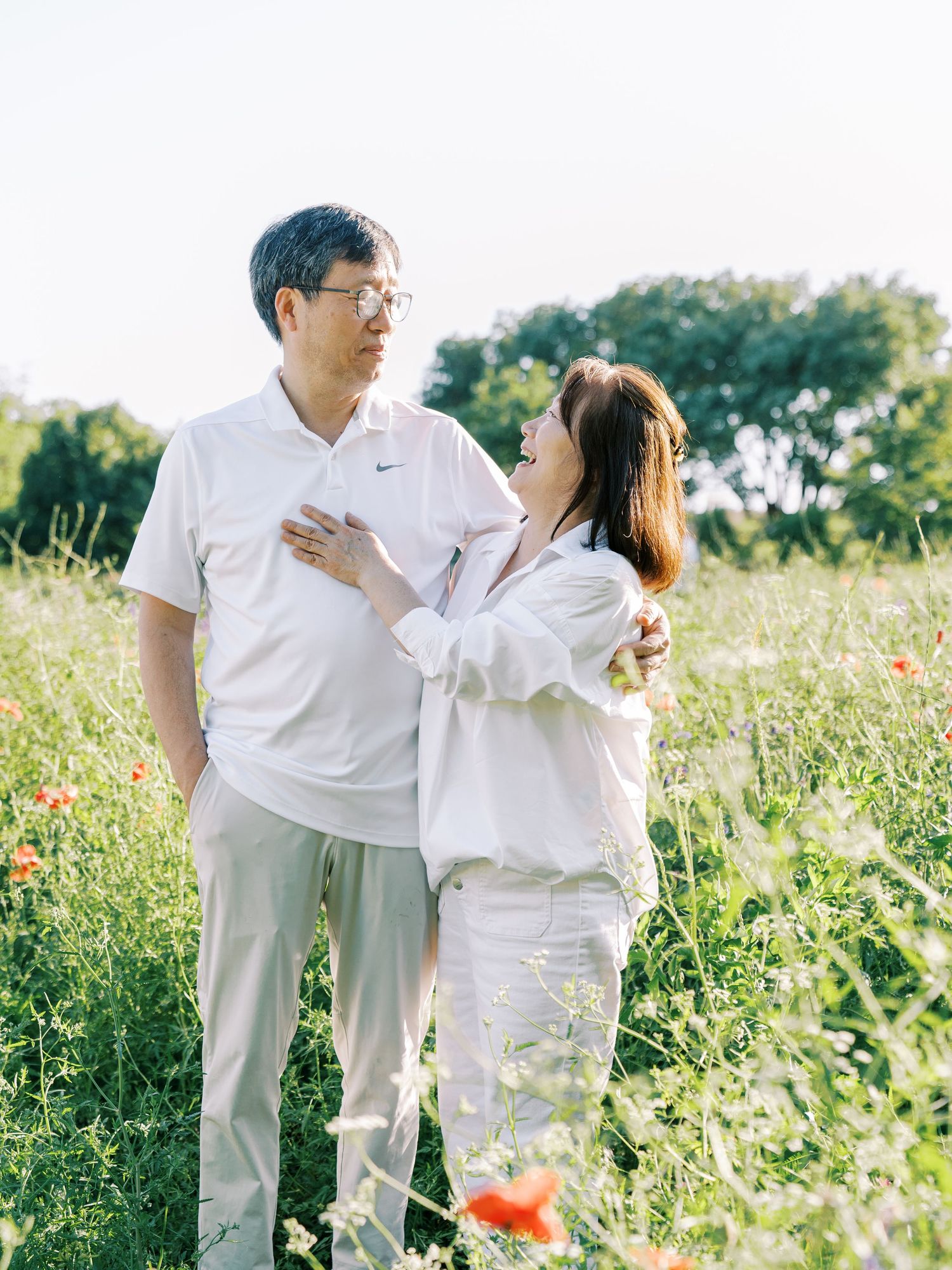 A couple in matching white outfits embraces while standing in a blooming summer meadow.