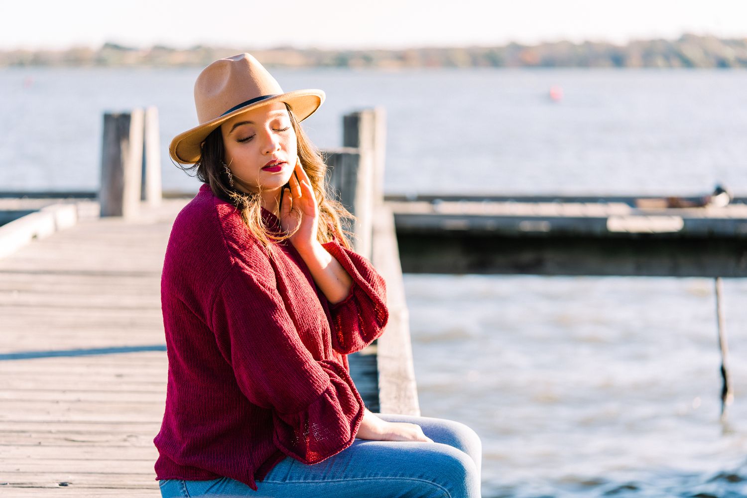 Person in red sweater and tan hat sits casually on wooden pier overlooking water.