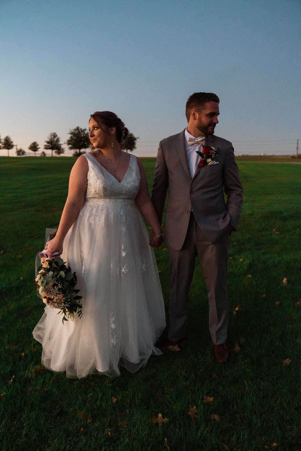 A couple in wedding attire walks through a green field at sunset, with the bride holding a floral bouquet.