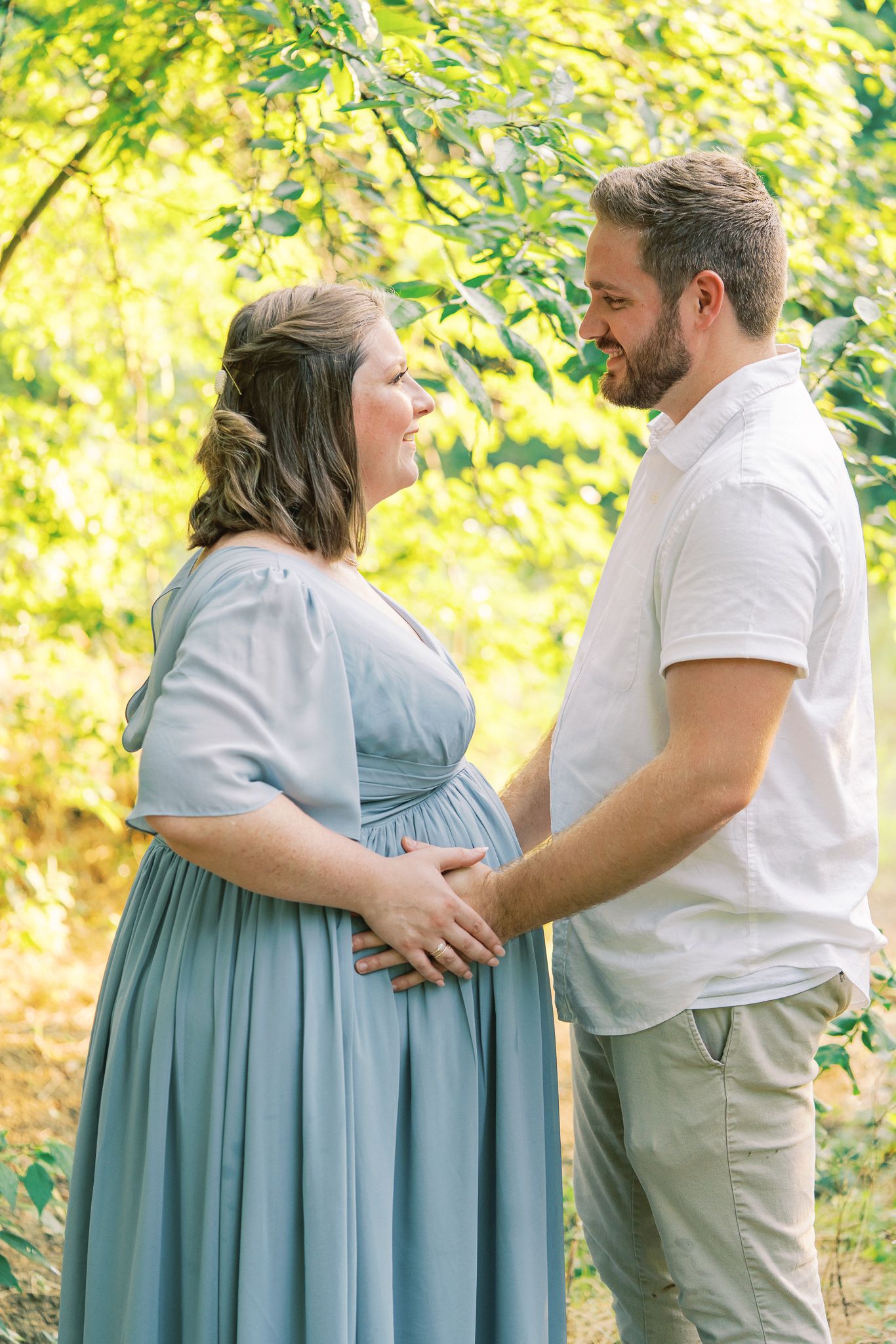 A couple in a natural setting share a tender moment as they celebrate an upcoming arrival in a light blue maternity dress.