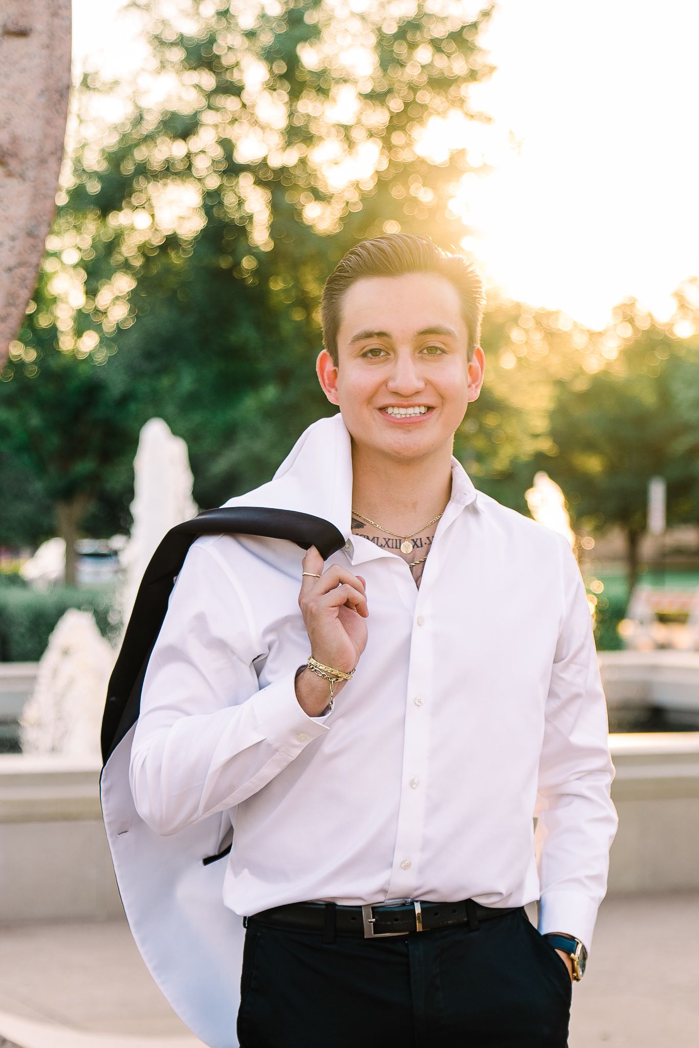 A person in a white dress shirt and black pants stands outdoors at sunset with a jacket casually draped over their shoulder.