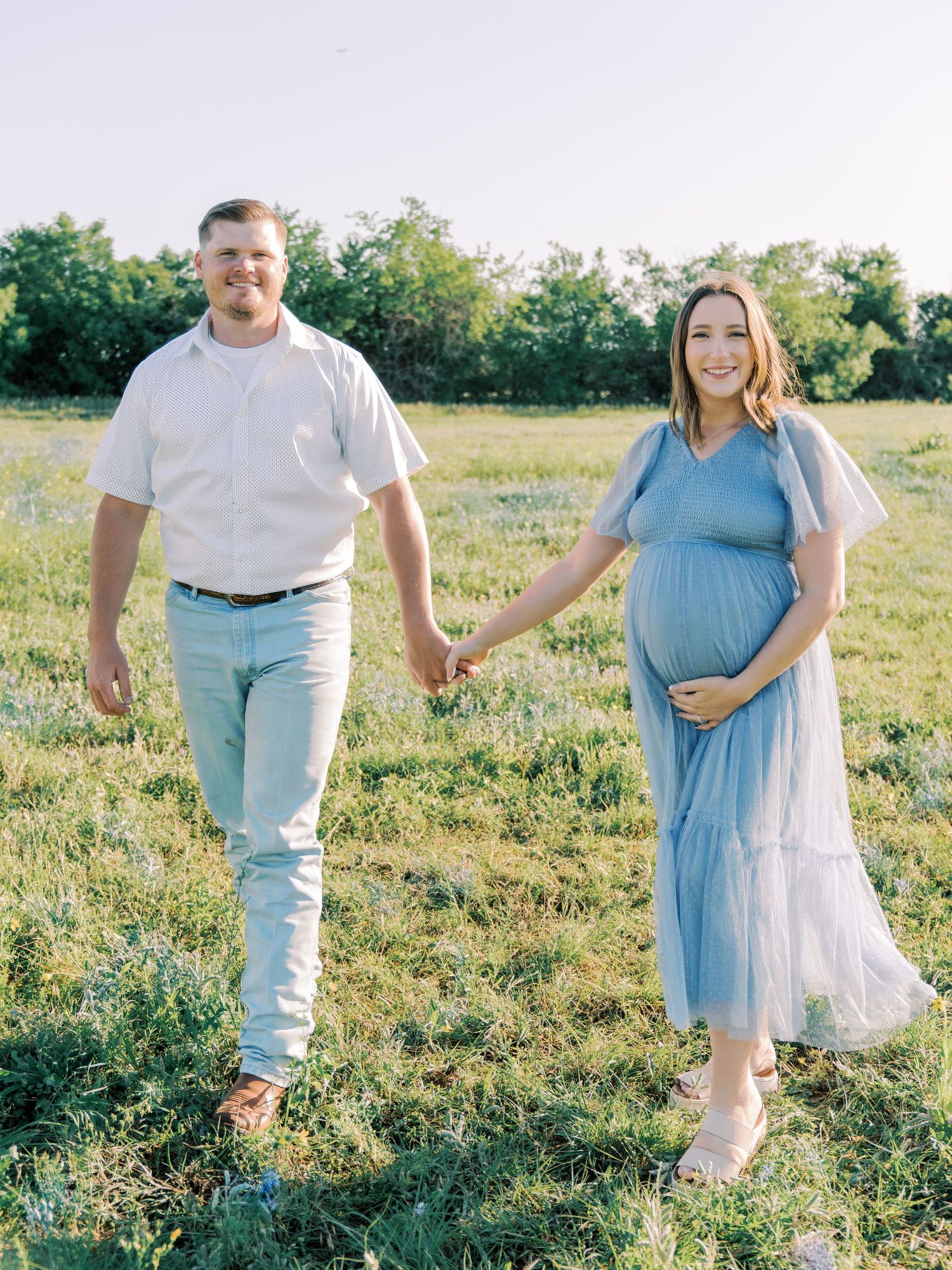 A couple in coordinating blue and white outfits poses together in a sunlit meadow during a romantic photoshoot.