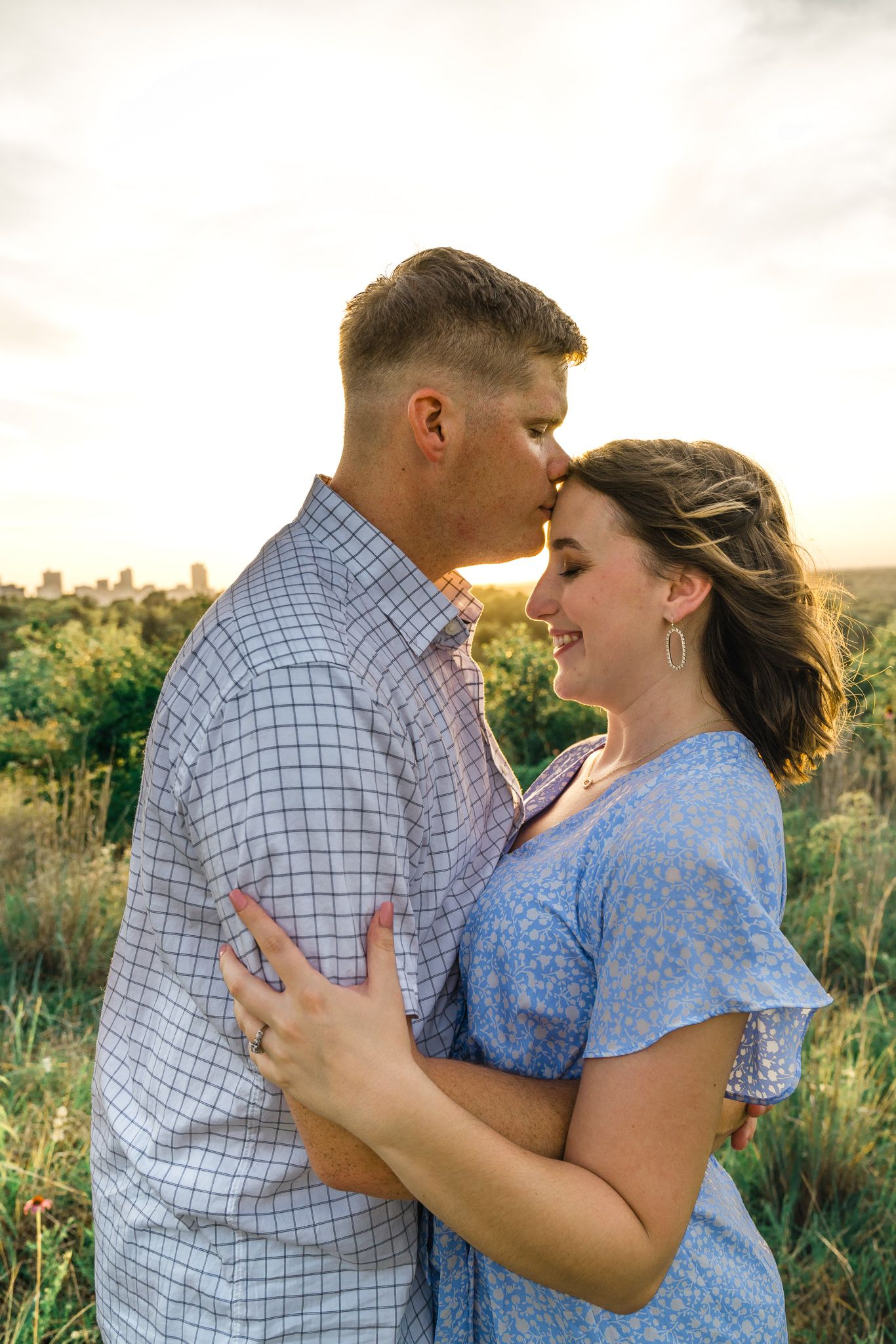 A couple shares a tender moment in a field during sunset, wearing blue and white patterned clothing.