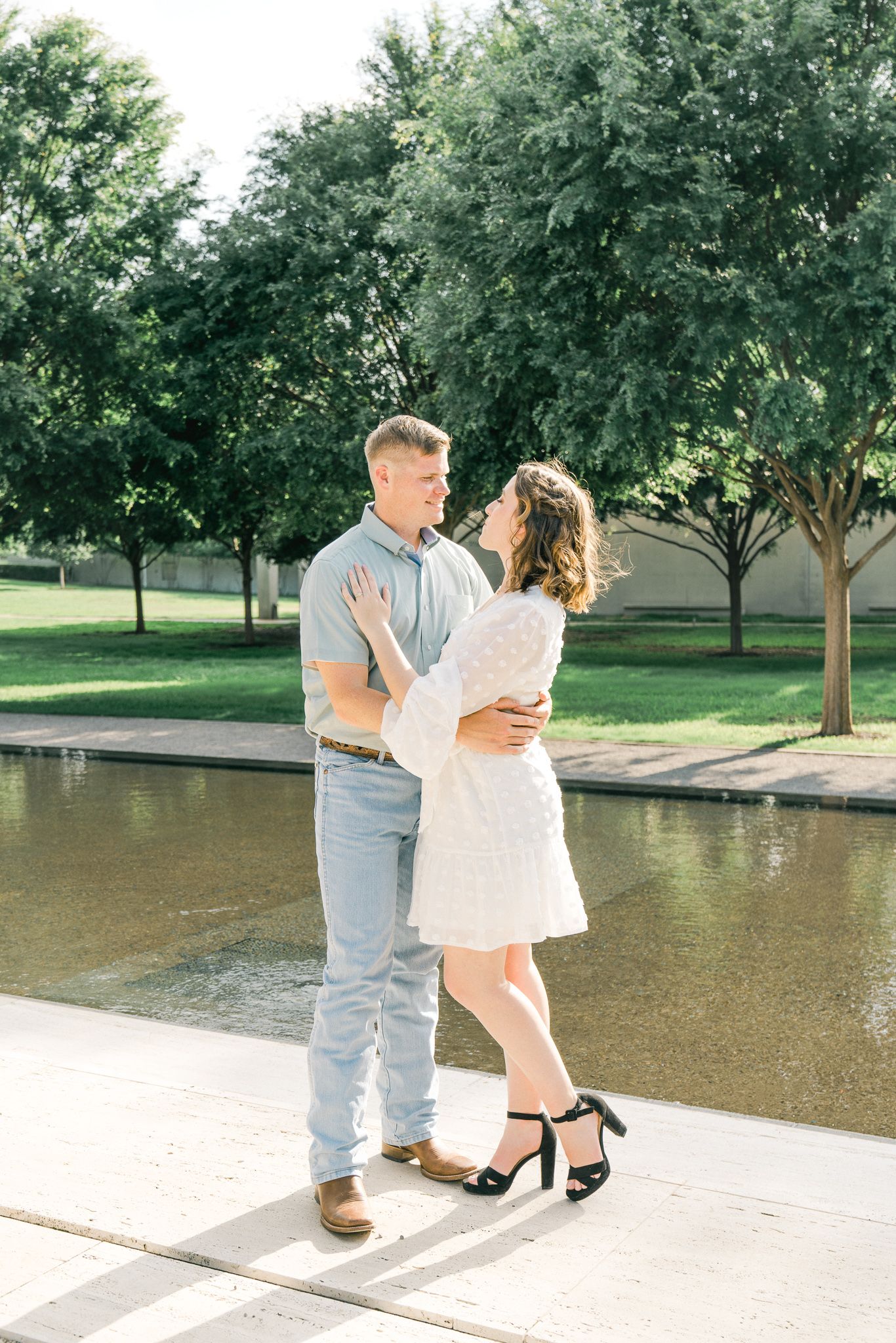 A romantic couple shares a tender moment while dancing together on a concrete path near a pond surrounded by green trees.