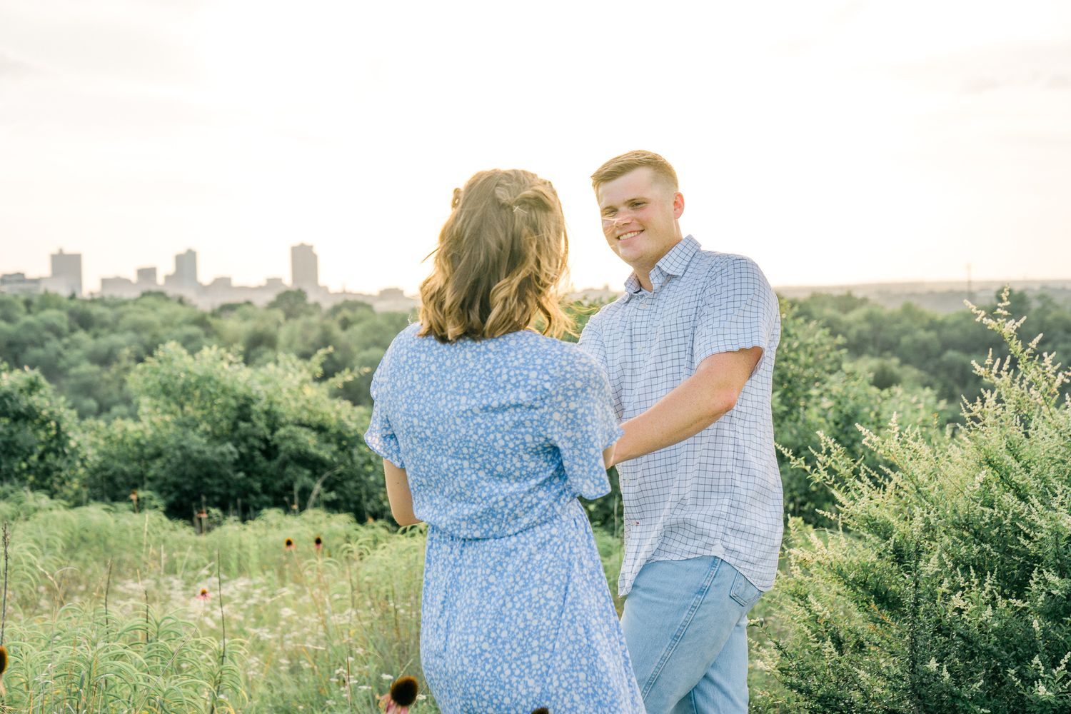 A couple shares a romantic moment in a wildflower field overlooking a distant cityscape.